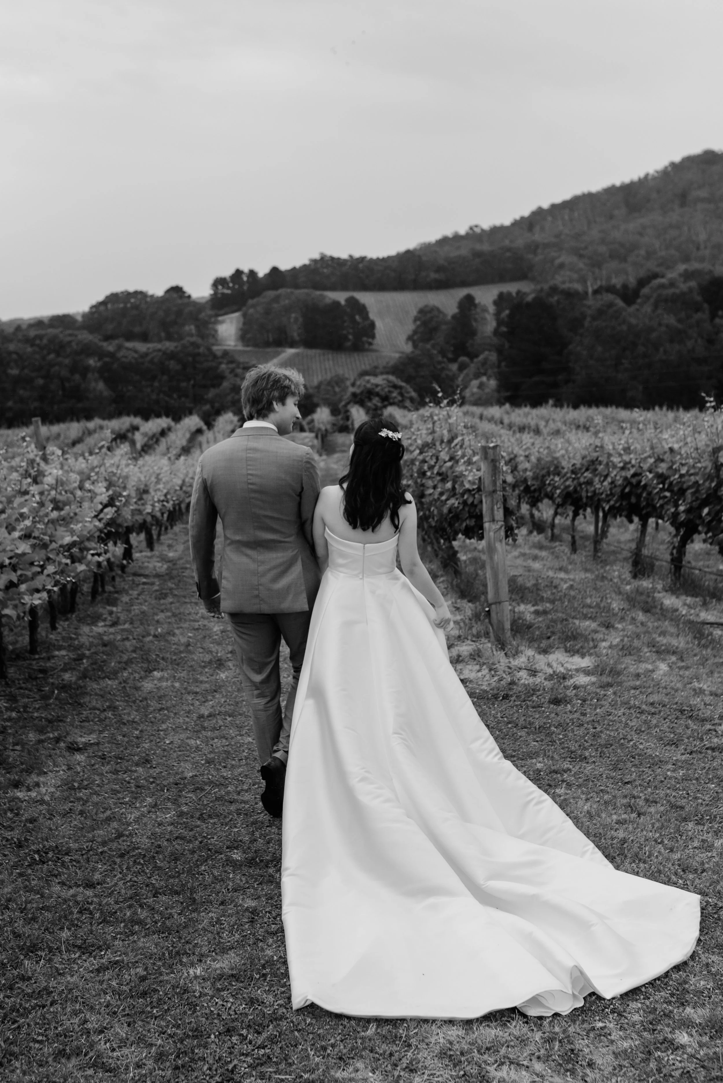 A couple in wedding attire walking through a vineyard, holding hands, with rolling hills in the background.
