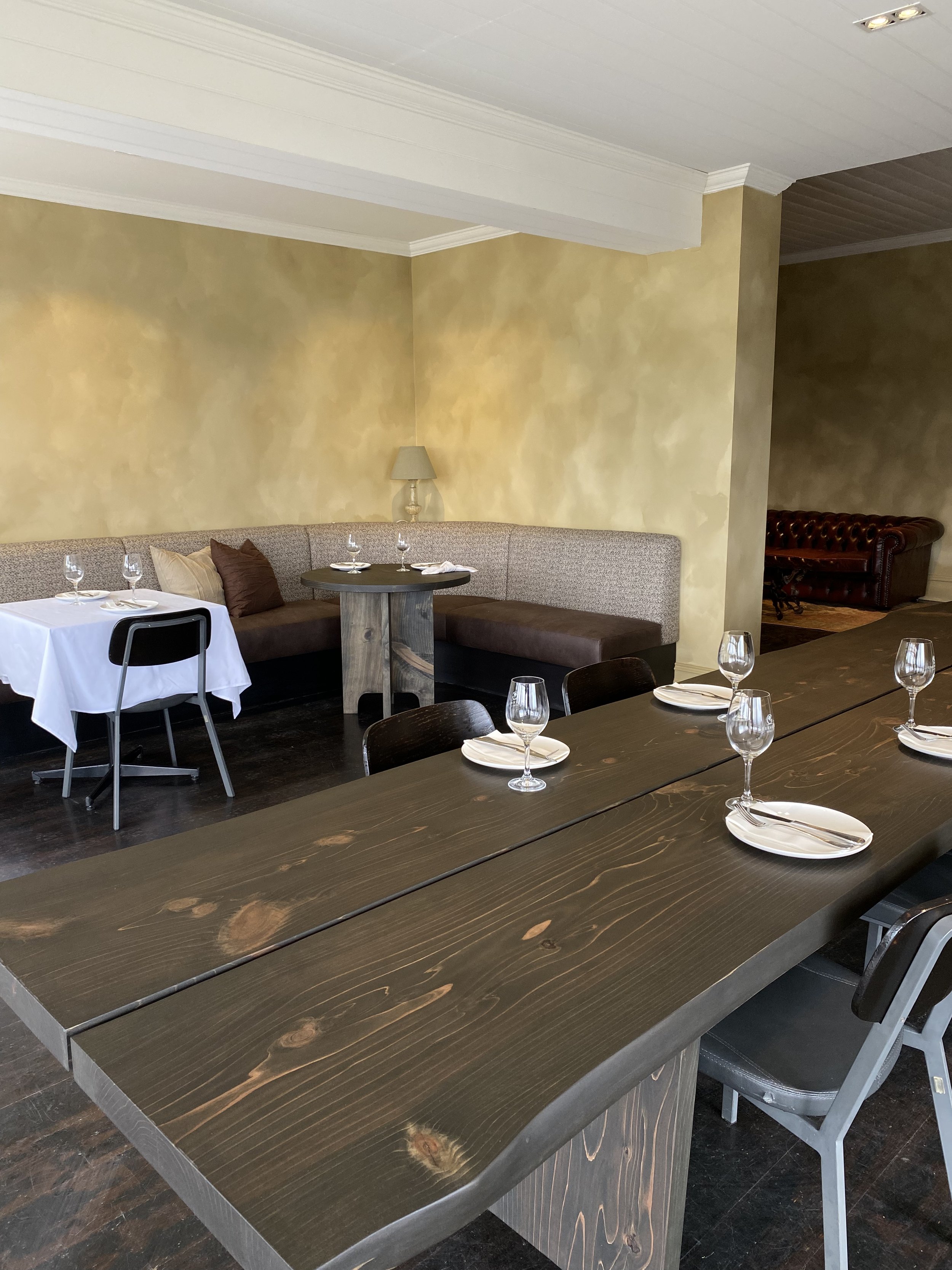 A restaurant interior with a large wooden dining table set with wine glasses, plates, and cutlery, a curved booth with pillows, and a corner lamp, with warmly painted walls and a dark wood floor.