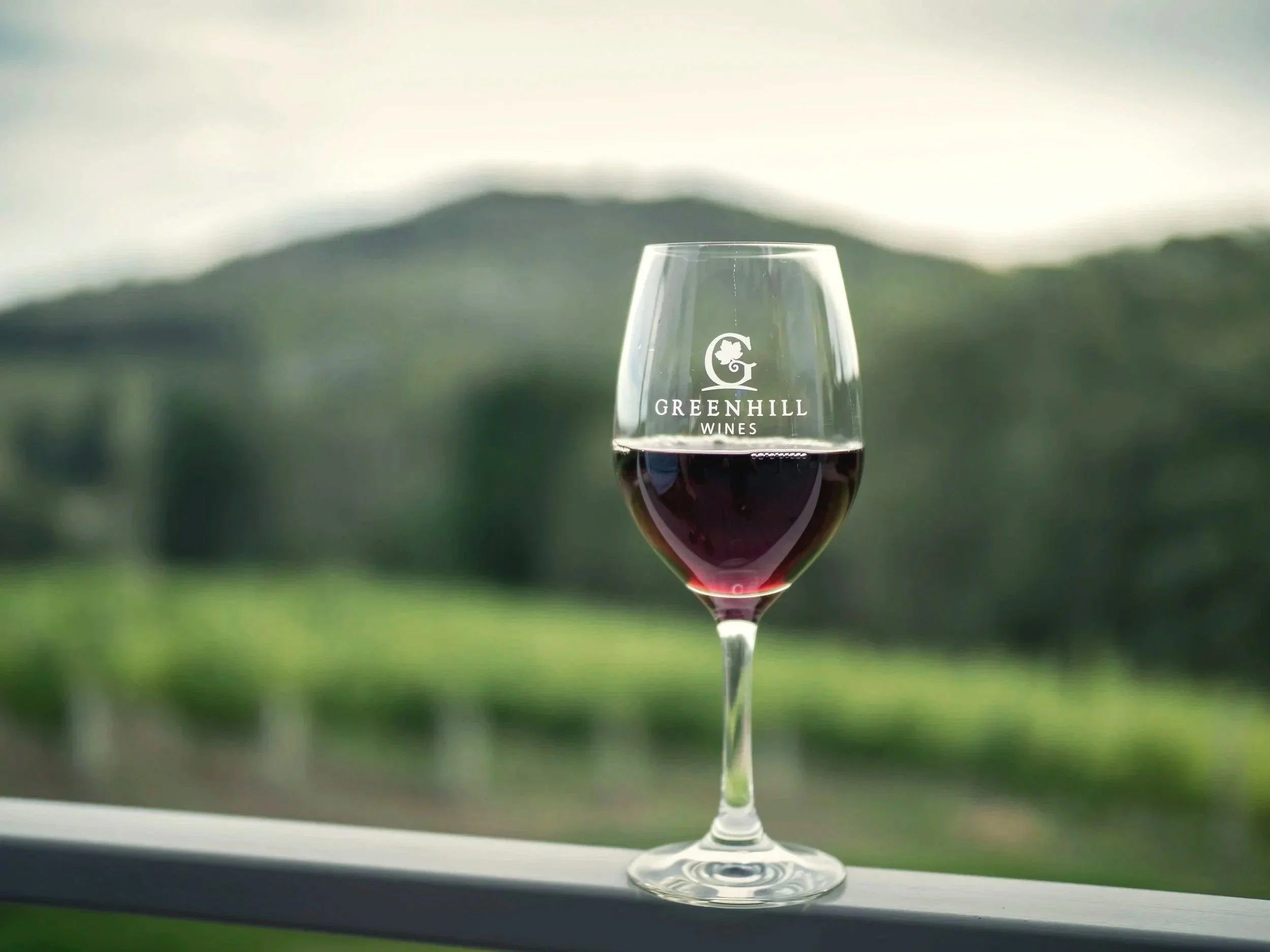 A wine glass with red wine, placed on a surface with a scenic blurred background of green hills and sky. The glass has the logo 'Greenhill Wines' on it.