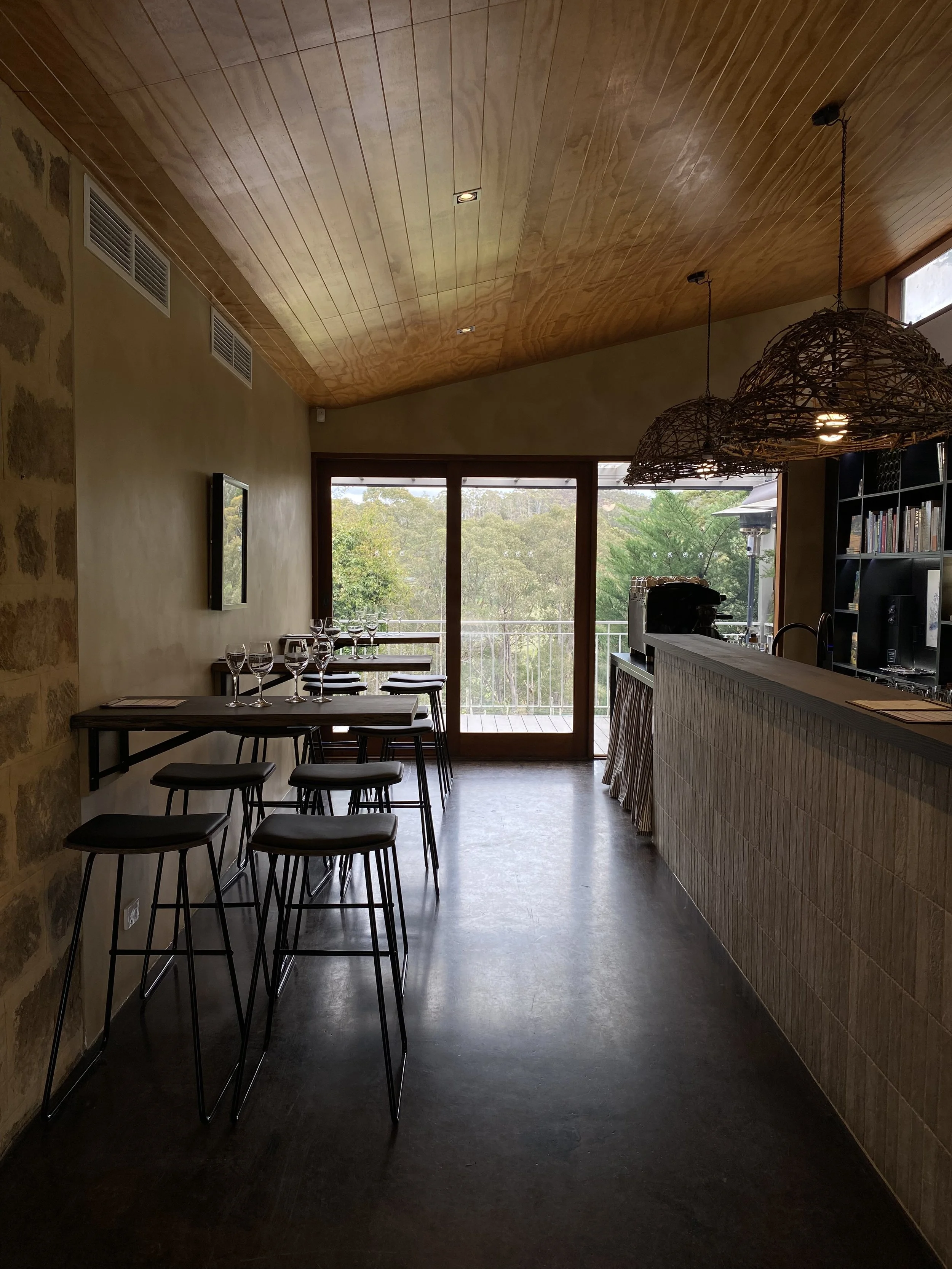 Interior view of a modern restaurant with dark flooring, a bar counter on the right, and large windows at the back overlooking trees. There's a small seating area with five bar stools and a set of glassware on the table. Wicker pendant lights hang from the wood-paneled ceiling.