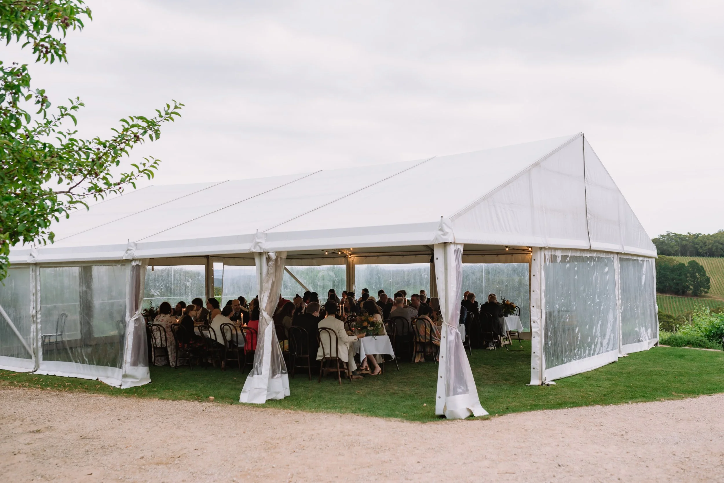 White outdoor event tent with transparent sides, filled with seated people at tables, set on grass with a gravel pathway outside, overlooking a green landscape with small hills and trees under a cloudy sky.