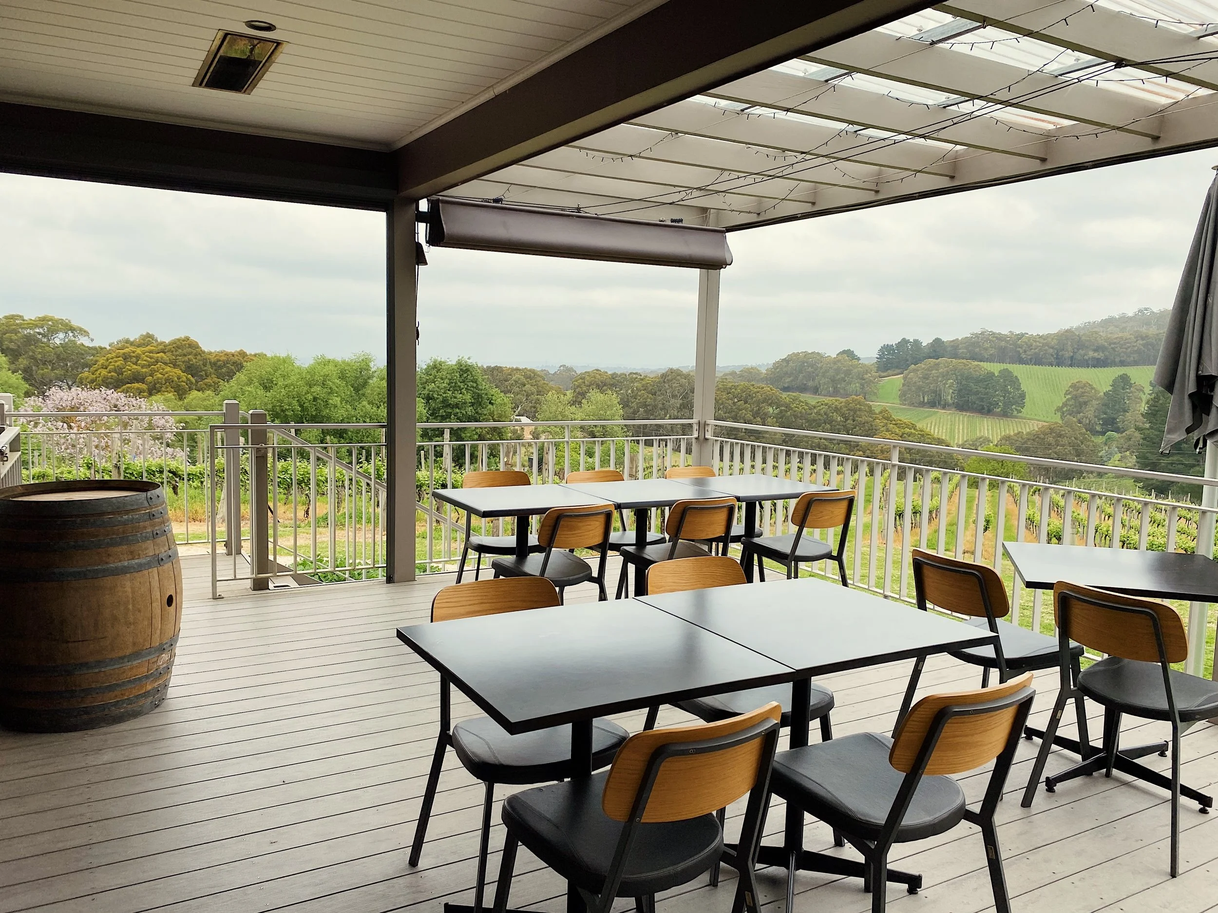 Outdoor balcony dining area with black tables, wooden chairs, a wine barrel, and a scenic view of rolling green hills, trees, and vineyards.