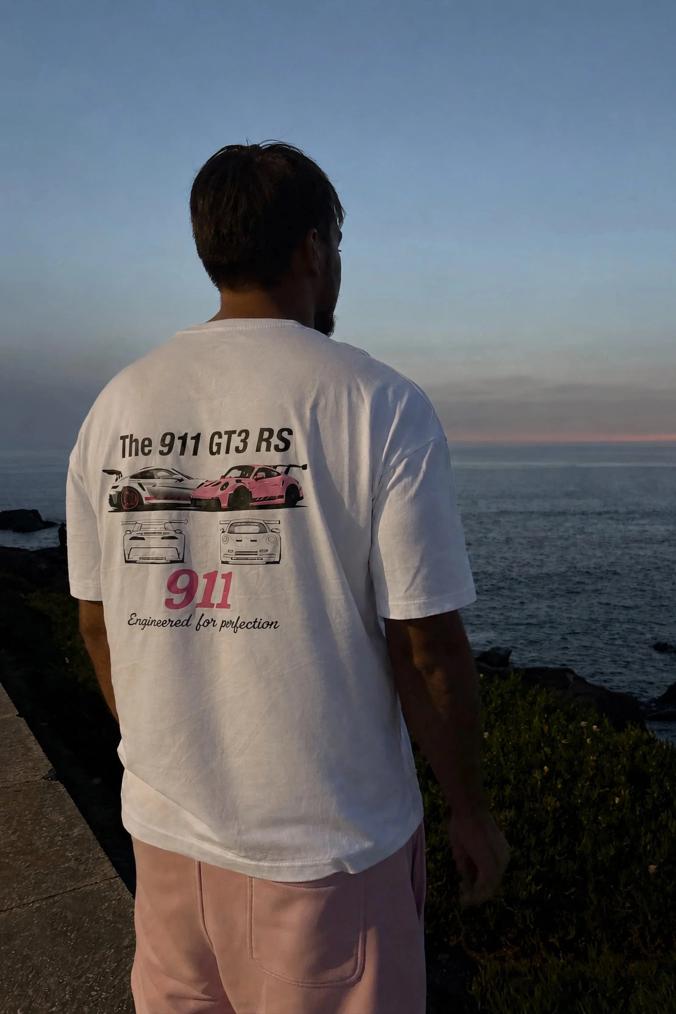Homme vu de dos regardant la mer au coucher du soleil, portant un t-shirt blanc avec des voitures de sport imprimées et le texte 'The 911 GT3 RS'.
