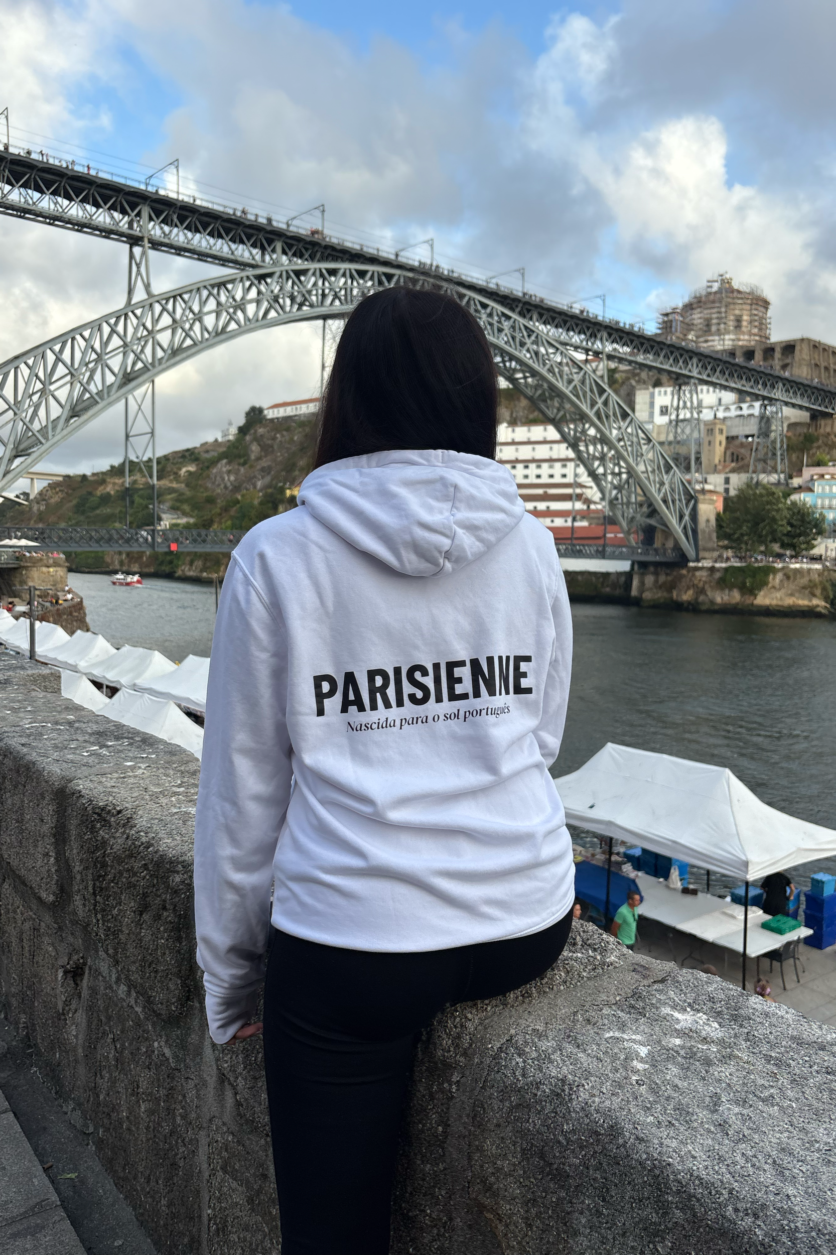 Femme assise sur un parapet, vue de dos, portant un sweat blanc avec le mot « Parisienne » écrit dessus, regardant le pont de Dom Luis à Porto, Portugal.