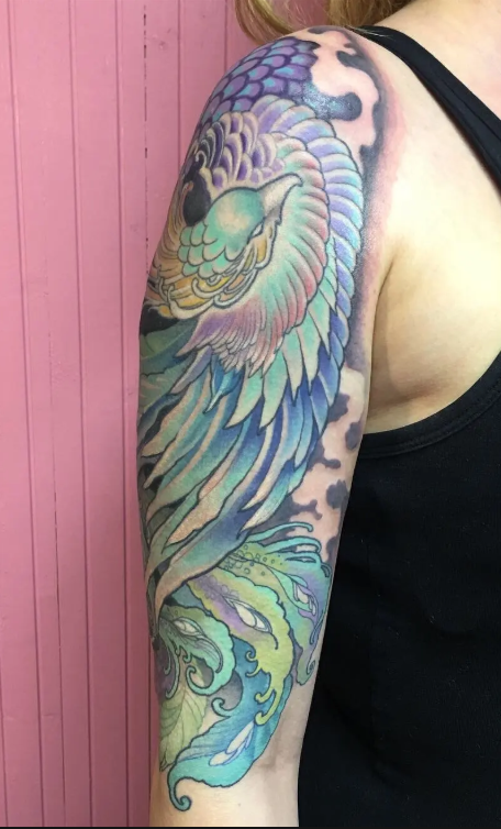 Colorful tattoo of a phoenix with wings, perched on a stylized swirl, on a person's upper arm and shoulder against a pink background.
