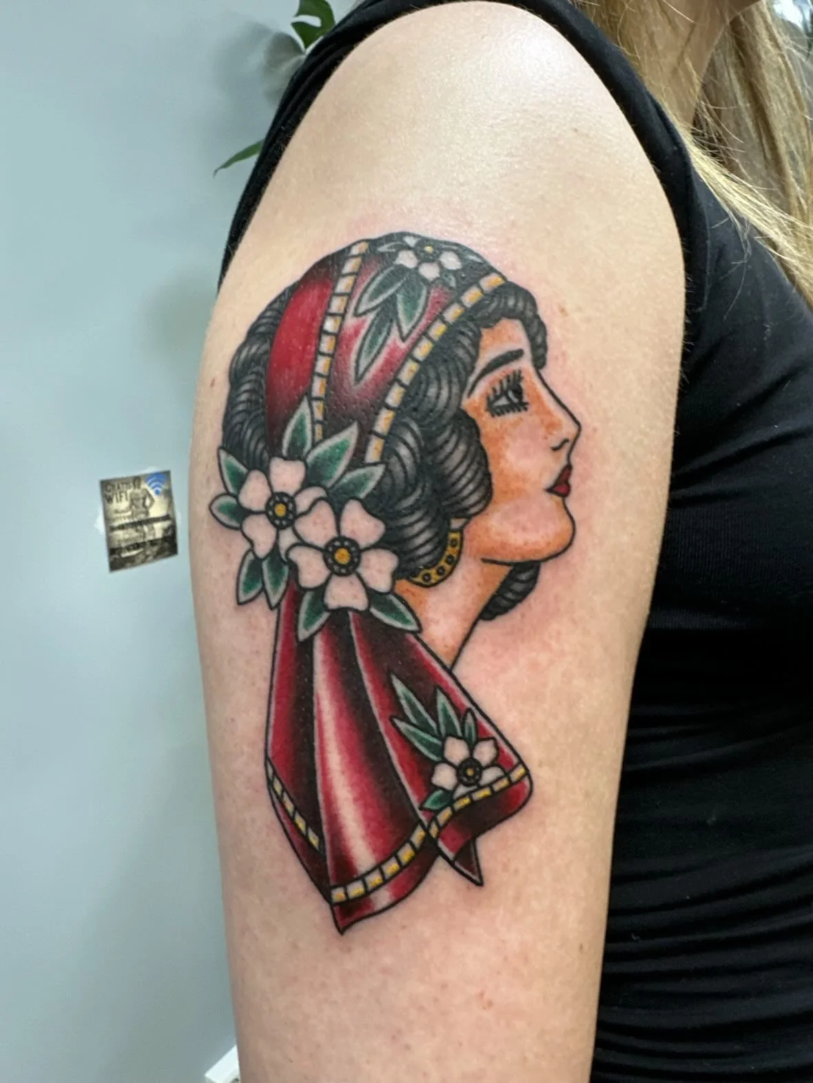 Tattoo of a woman in profile with dark, curly hair, wearing a red headscarf and flowers, on upper arm.