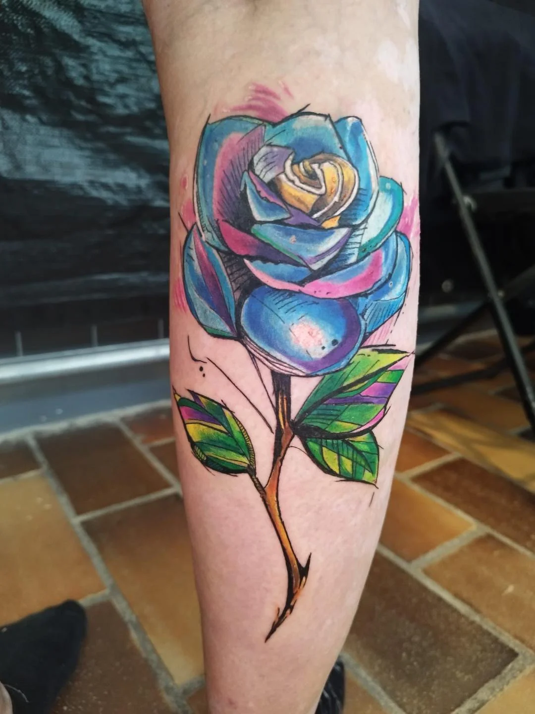 Colorful watercolor-style tattoo of a blue and purple rose with green leaves on a person's leg.