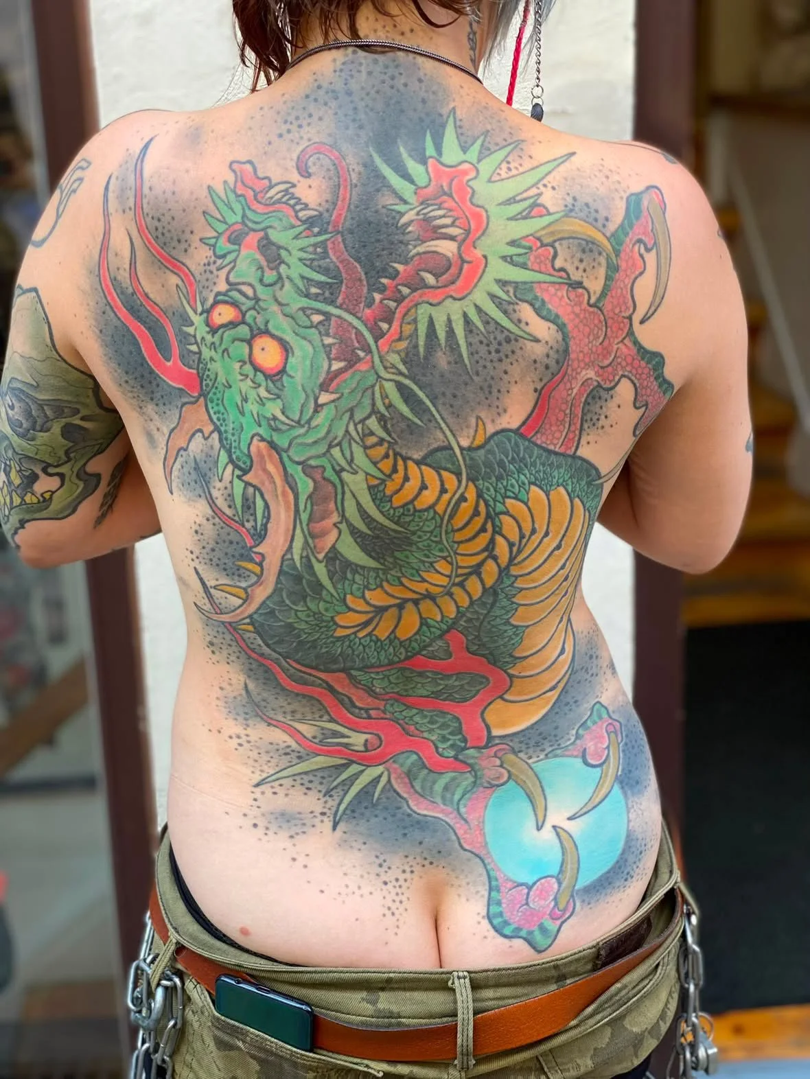 Colorful tattoo of a dragon on a person's back, with green, orange, and red hues, detailed scales, and a fierce expression.