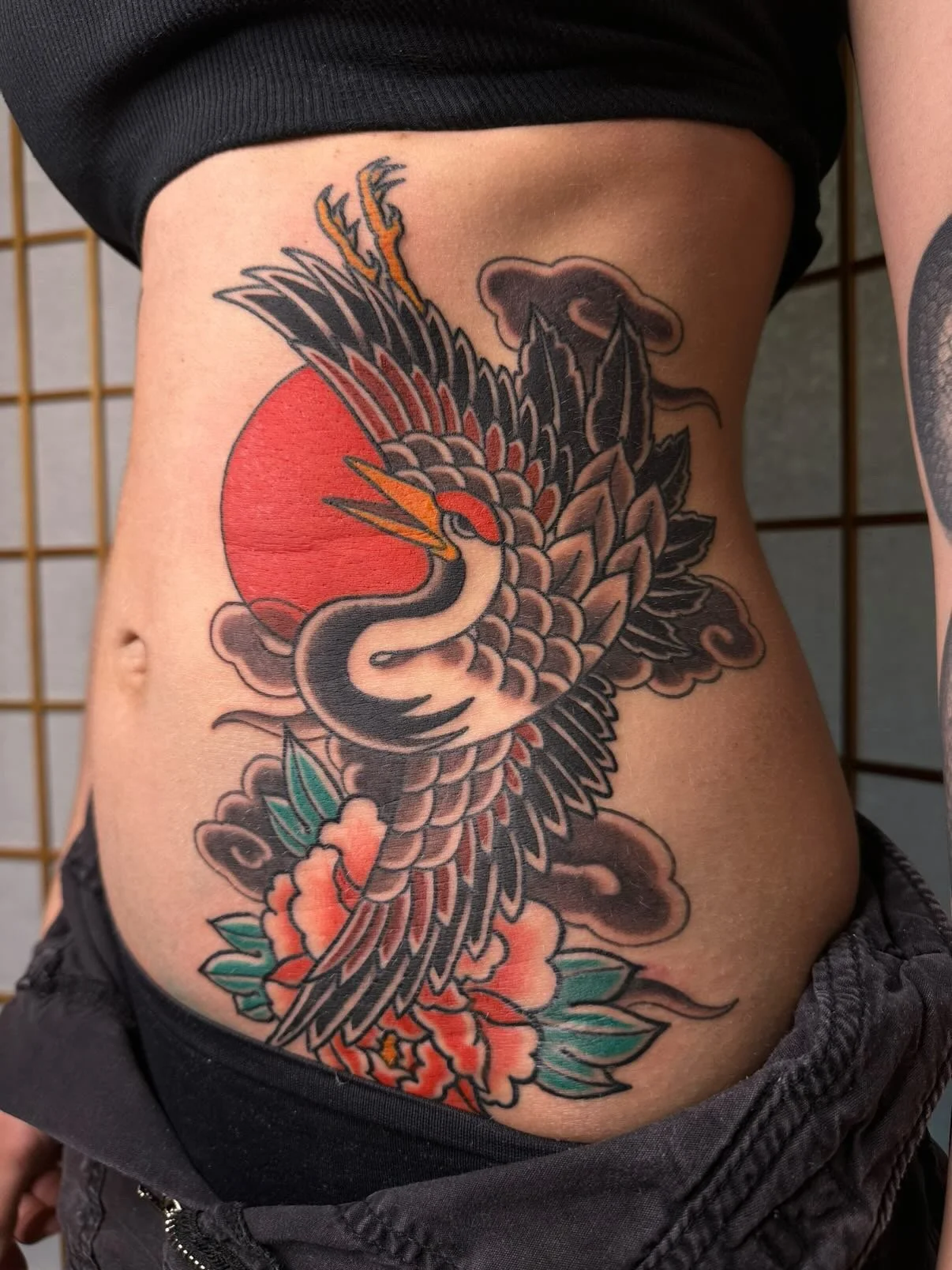 Tattoo of a Japanese-style crane with wings spread, flying in front of a red sun, with colorful flowers and green leaves below.