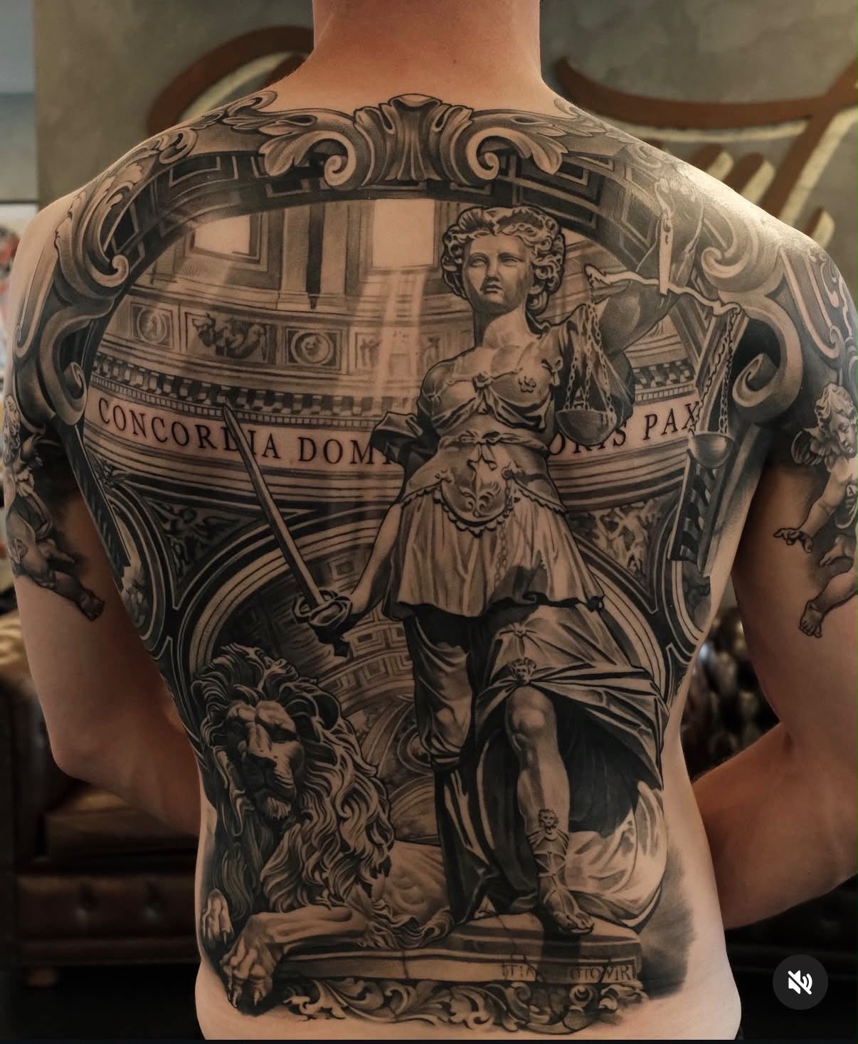 Black and gray tattoo of a classical statue of a woman holding a sword and shield, surrounded by intricate architectural and ornamental elements, including a lion's head at the bottom and Latin inscriptions.