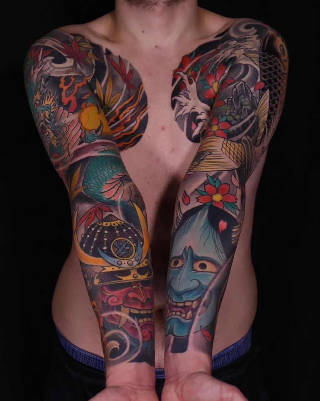 A shirtless person with extensive, colorful traditional Japanese-style tattoos covering their arms, chest, and shoulders, featuring dragons, flowers, masks, and other classic motifs.