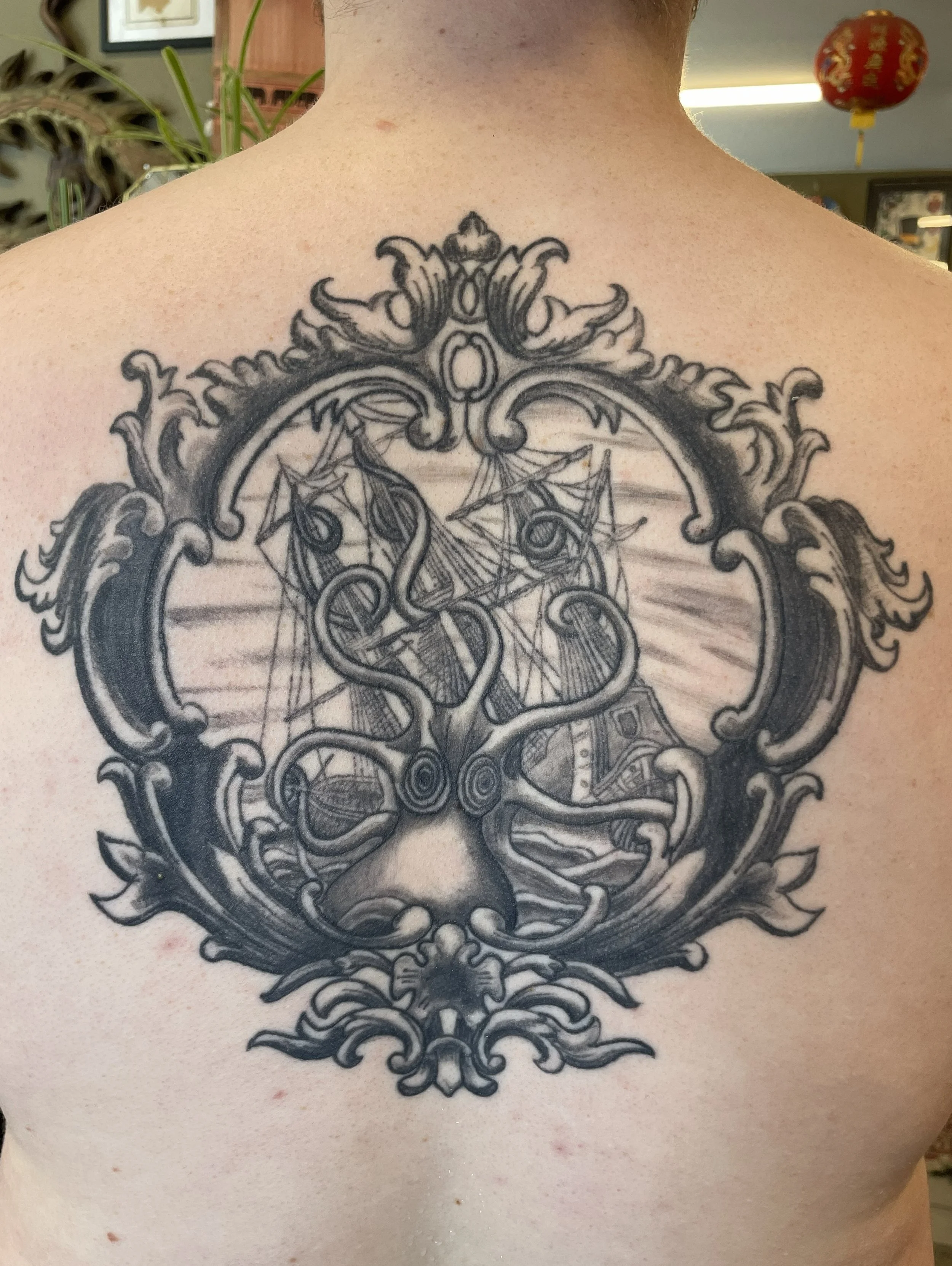 A black and gray tattoo on a person's upper chest and neck depicting a complex, ornate design with symmetrical swirling elements and detailed line work.