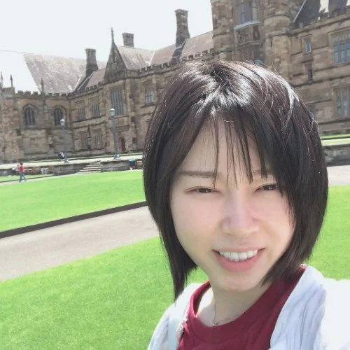 A young woman with short black hair smiling outdoors in front of a historic stone building with a well-maintained green lawn.
