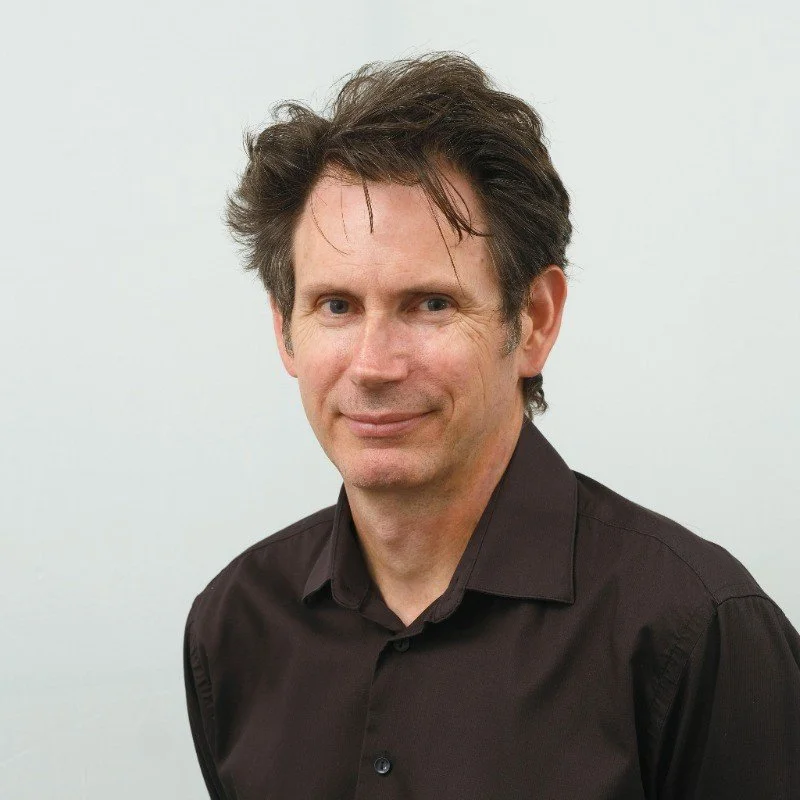 Headshot of a middle-aged man with dark brown, slightly wavy hair, wearing a black collared shirt, smiling slightly, against a light background.