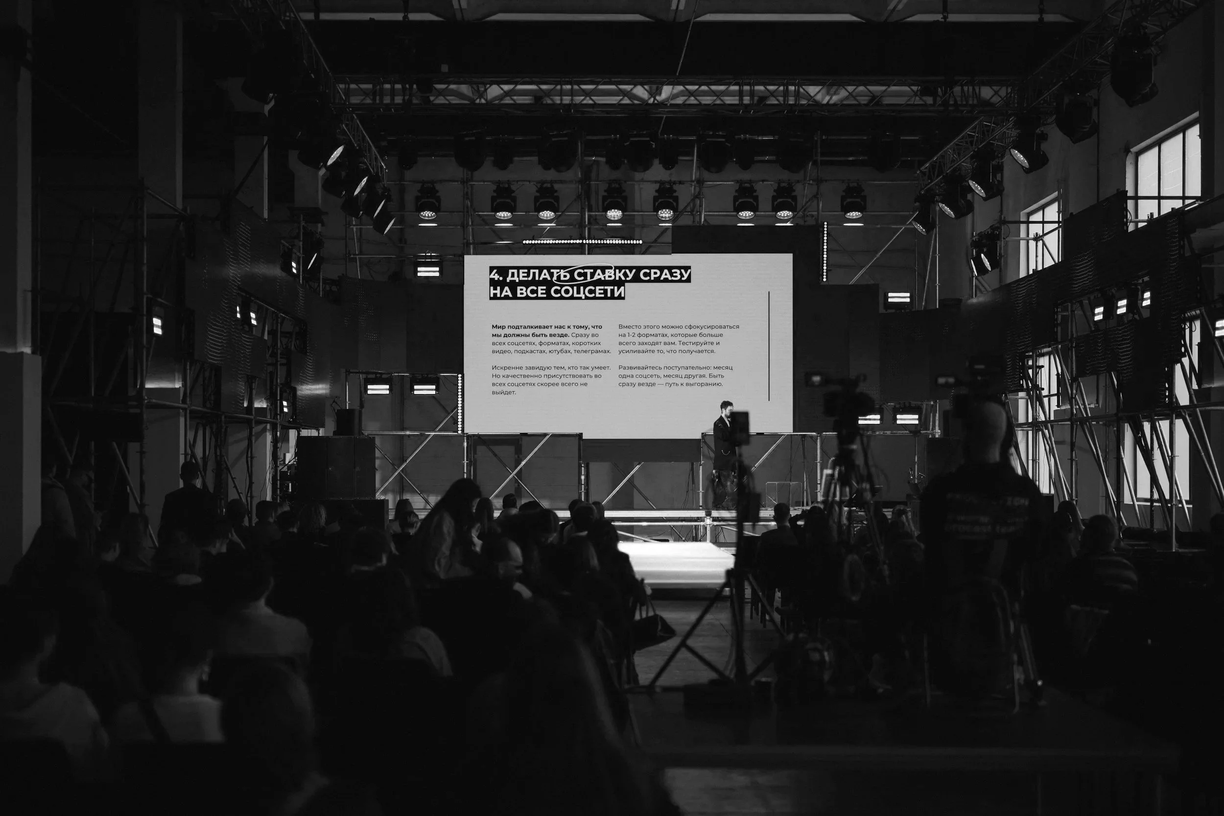 A conference hall with a stage and a large screen displaying a presentation in Russian, an audience seated in front, and a speaker standing on the stage. The room has high ceilings and large windows on the side.