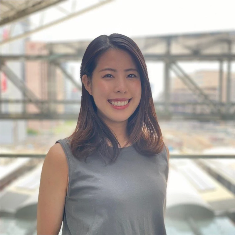 A woman with shoulder-length dark hair smiling while standing in front of a blurred cityscape background.