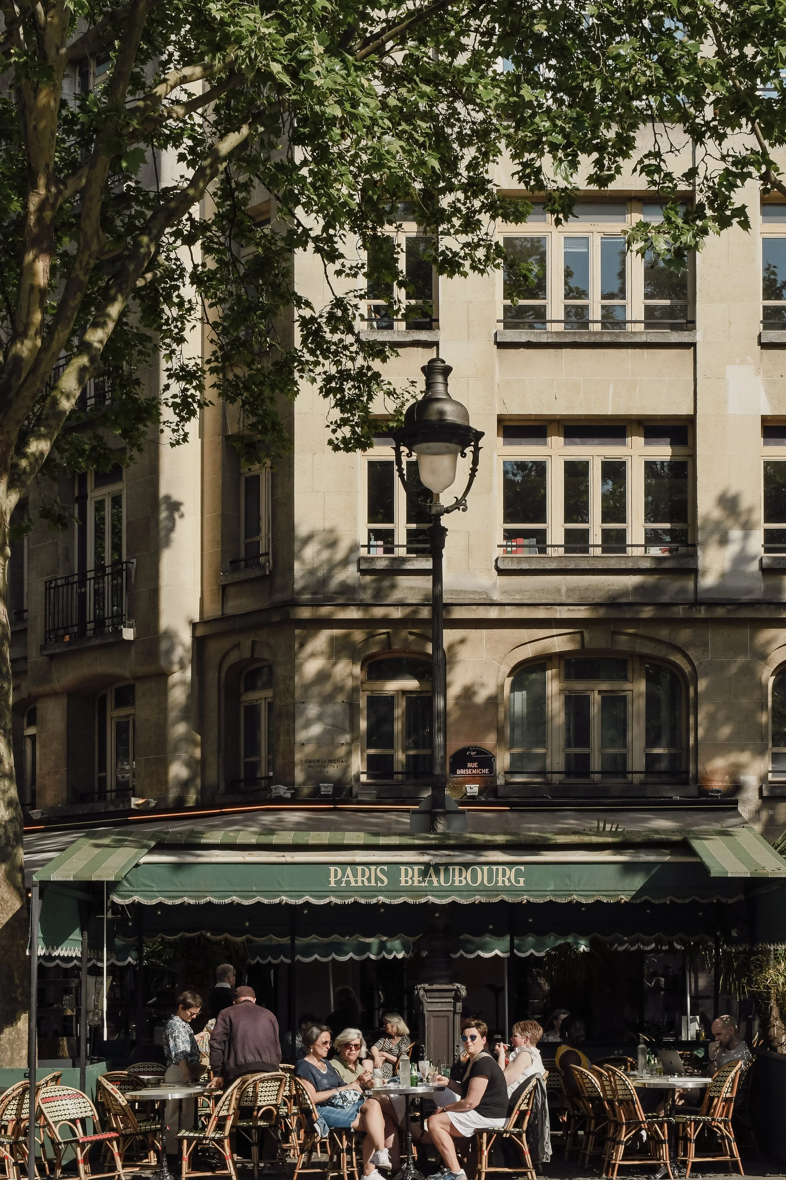 Terrasse de café à Paris avec des clients assis, un parasol marqué 'PARIS BEAUBOURG', un bâtiment en arrière-plan avec de grandes fenêtres et un lampadaire antique, ombragé par un arbre.