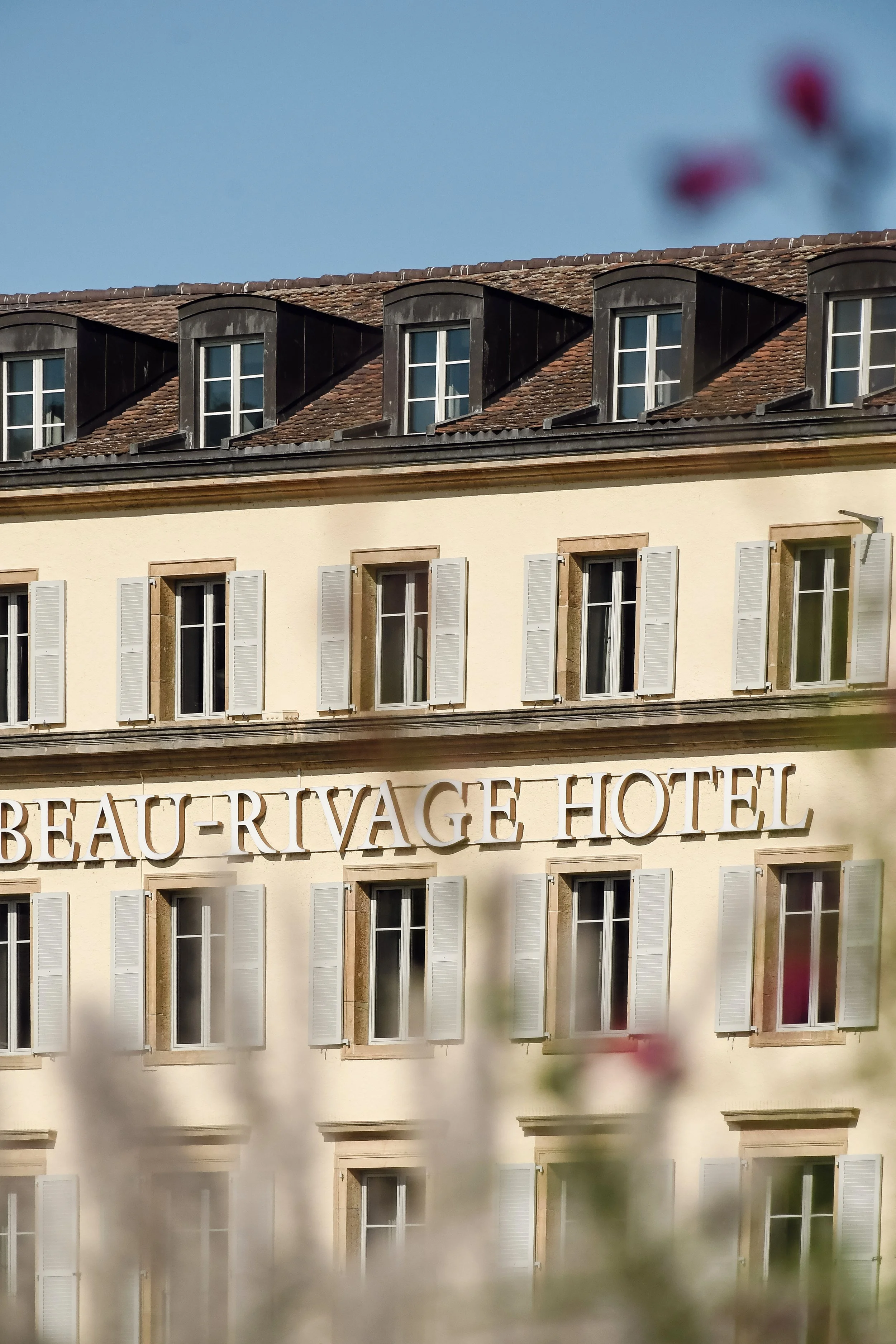 Facade d'un hôtel nommé 'Beau-Rivage' avec plusieurs fenêtres à volets blancs, un toit en ardoise avec des lucarnes et des fenêtres à double vitrage.