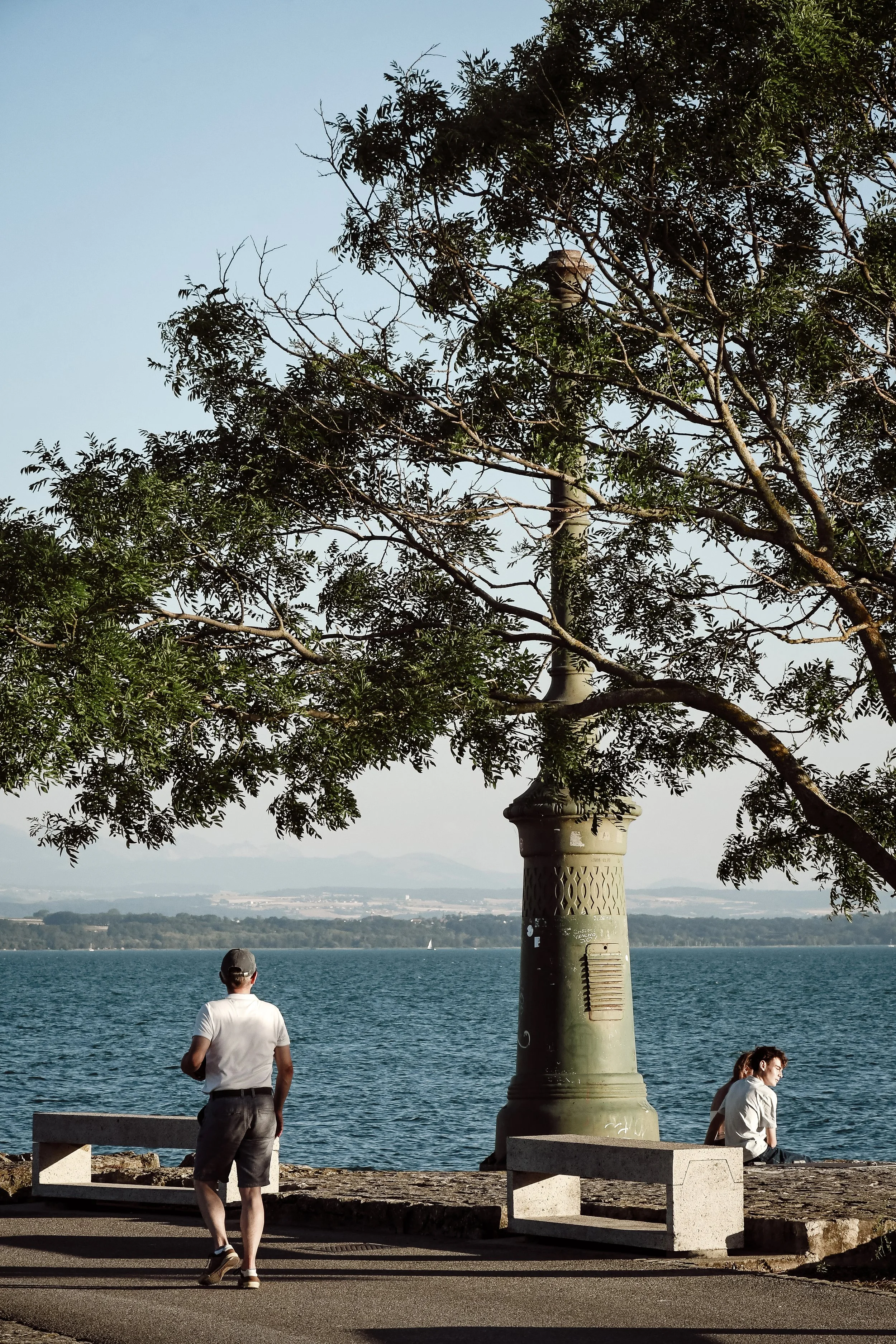 Un homme vêtu d'une chemise blanche et d'un pantalon noir se tient face à un lac, avec une structure en pierre à sa gauche et deux autres personnes assises sur un banc à droite. Un arbre avec des feuilles vertes s'élève derrière la structure en pierr