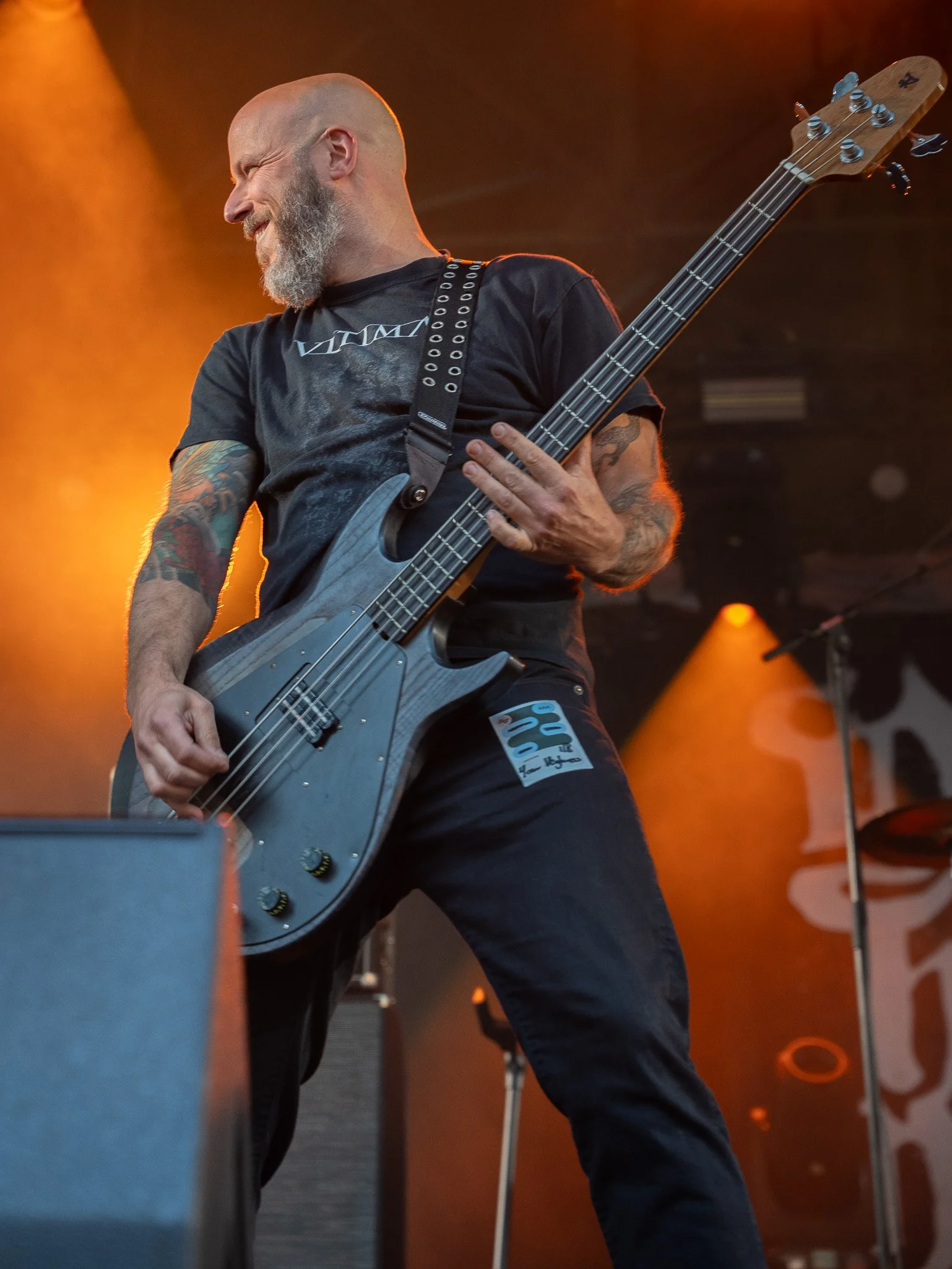 A male musician playing bass guitar on stage with orange lighting behind him, wearing a black T-shirt and black pants, with tattoos on his arms and a gray beard.