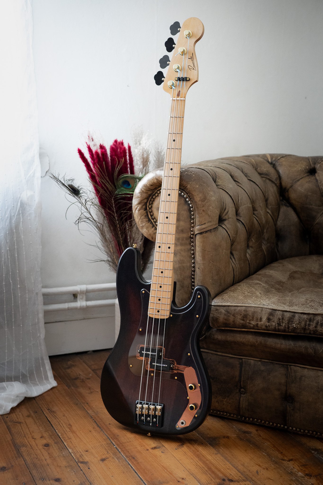 Een zwarte elektrische basgitaar met een houtkleurig toets en gouden hardware staat rechtop in een kamer, naast een oude leren bank en een vaas met rode en bruine pluimen. Aan de muur hangen zwarte muurdecoraties.