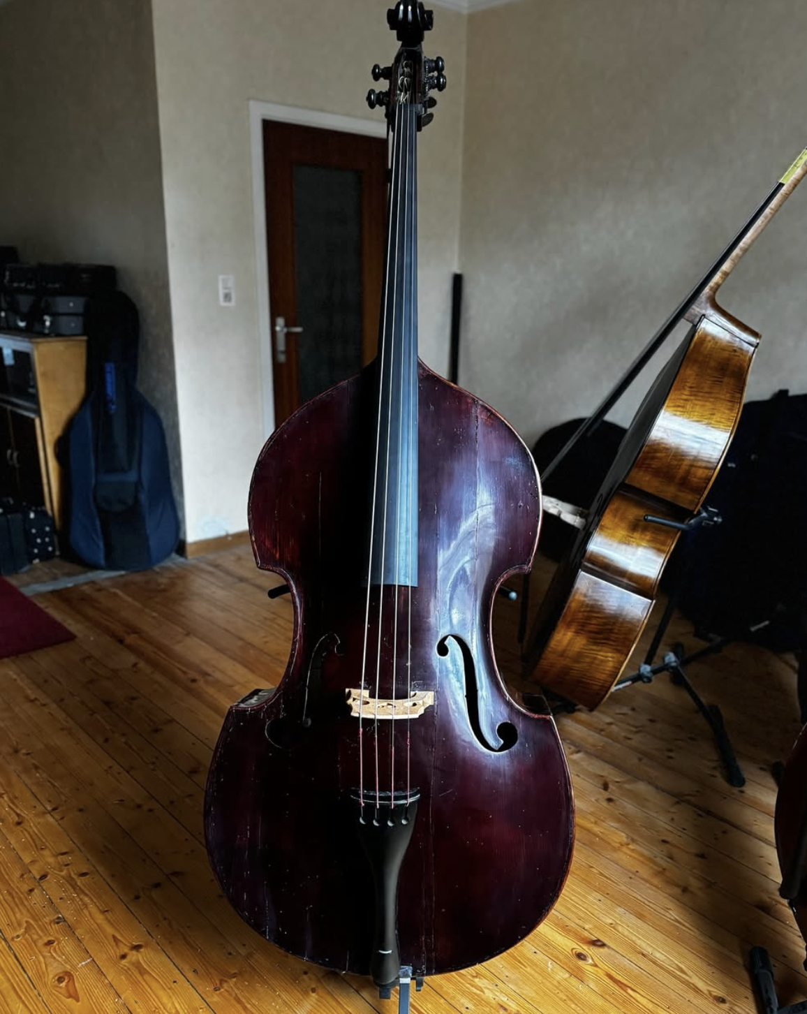 An upright double bass in the foreground with a wooden acoustic guitar in the background inside a room with wooden floors.