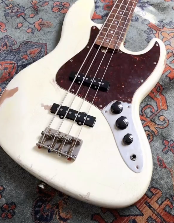 A vintage white electric bass guitar with a dark tortoiseshell pickguard and three control knobs, resting on a patterned rug.