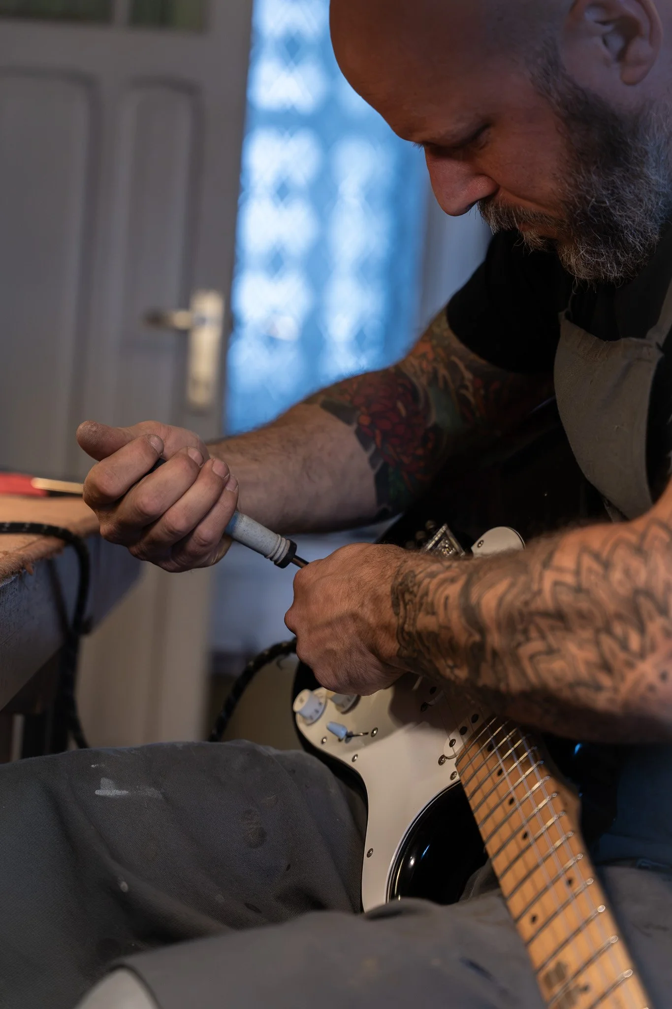 Man met bearmasker en tattoos die een elektrische soldeerbout vasthoudt en aan een elektrische gitaar werkt, binnen in een kamer.