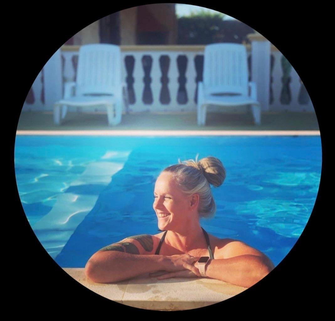 Vrouw met een vacht, blonde haar in een knot, ontspannen in een zwembad, lachend, met een zwembad en ligstoelen op de achtergrond
