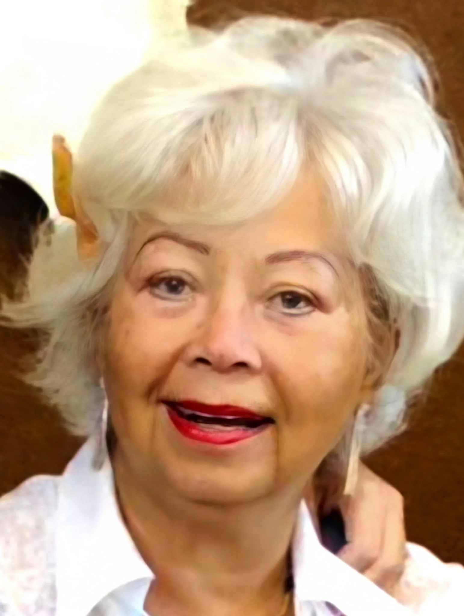 Close-up van een glimlachende oudere vrouw met wit haar, rode lippenstift en lichte huid, met een persoon op de achtergrond.