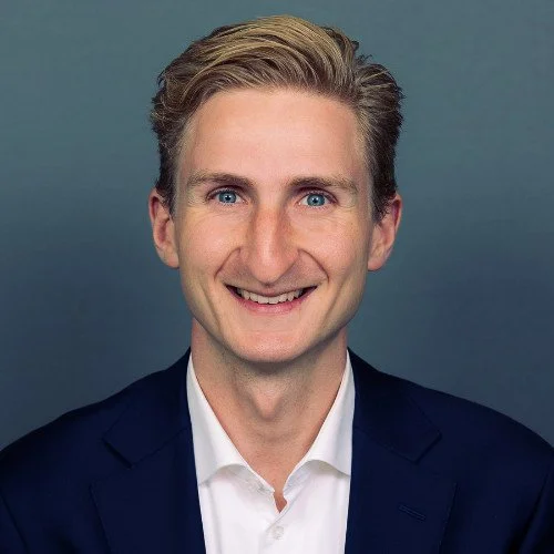 Portret van een man met blond haar, blauwe ogen, glimlach, in een donkerblauwe jas en witte blouse, tegen een grijze achtergrond.