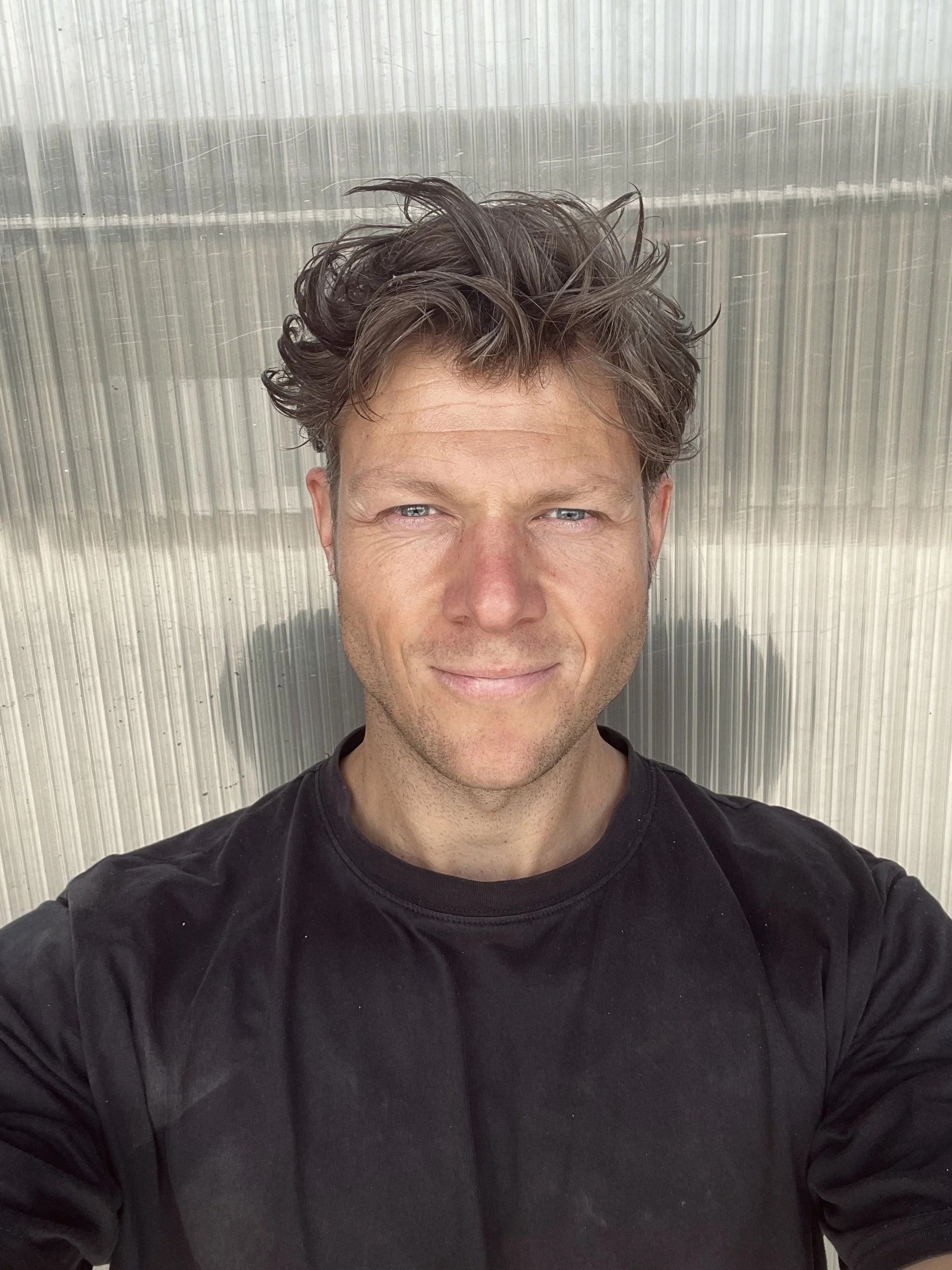 Een man met bruin, wave-achtig haar en blauwe ogen die een zwarte T-shirt draagt. Op de achtergrond is een glazen muur te zien.