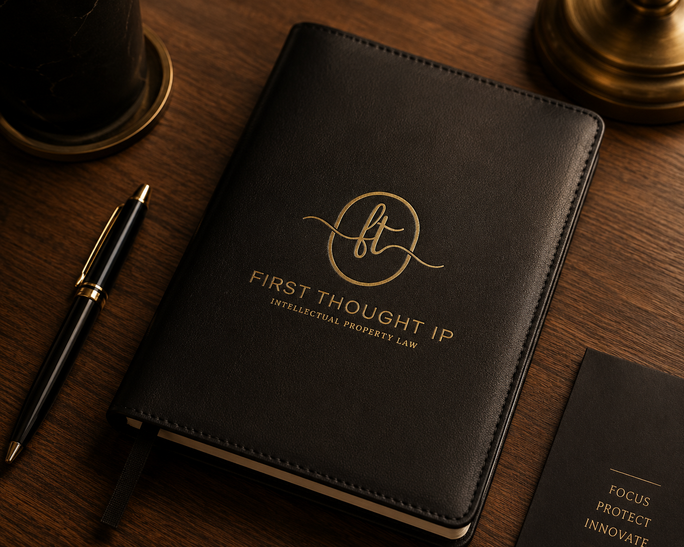 Black leather notebook with gold lettering and logo, alongside a black and gold pen on a wooden desk.