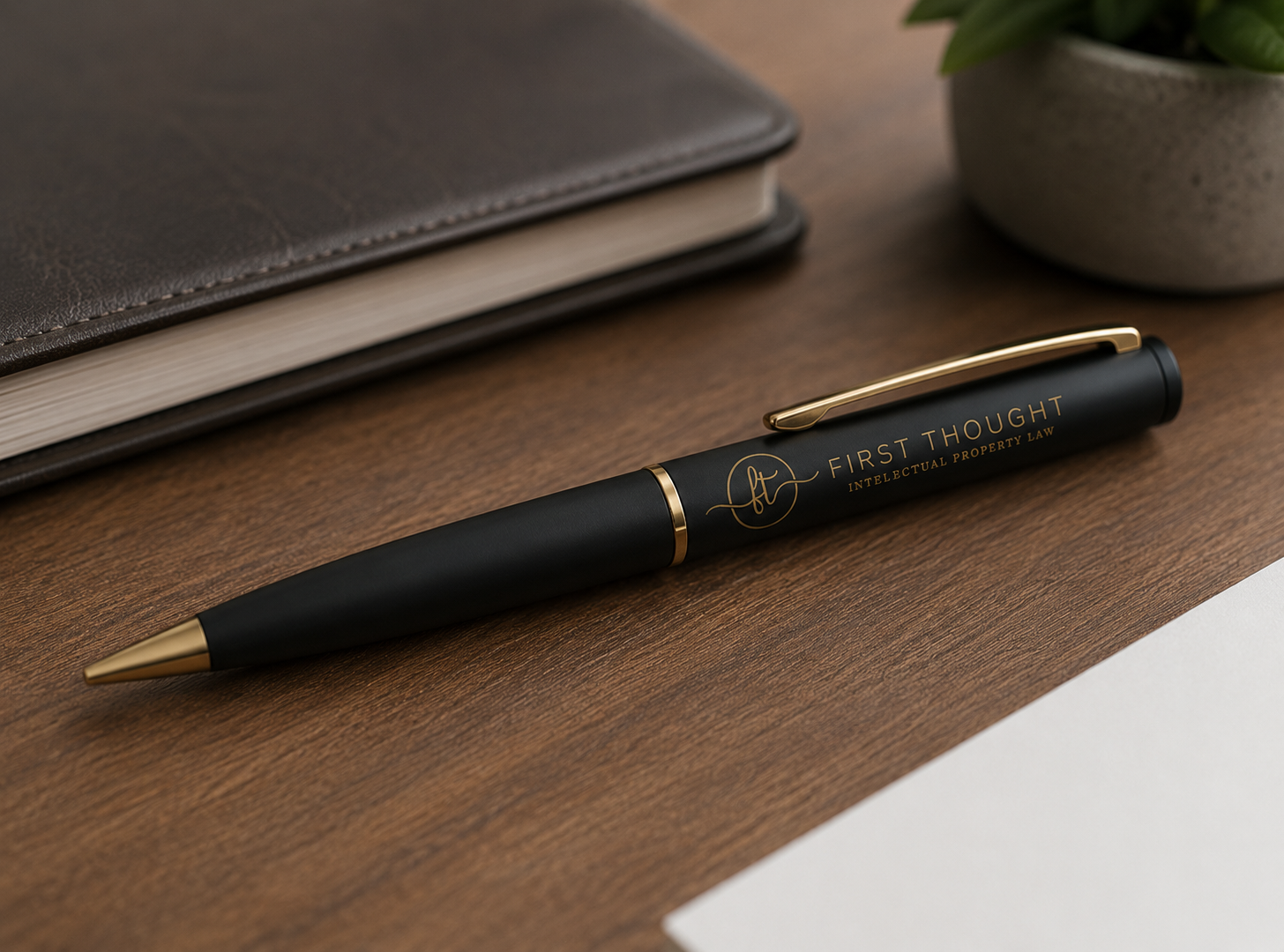 Black pen with gold accents reading "First Thought" and "Intellectual Property Law" on a wooden desk, near a closed brown leather notebook and a potted green plant.
