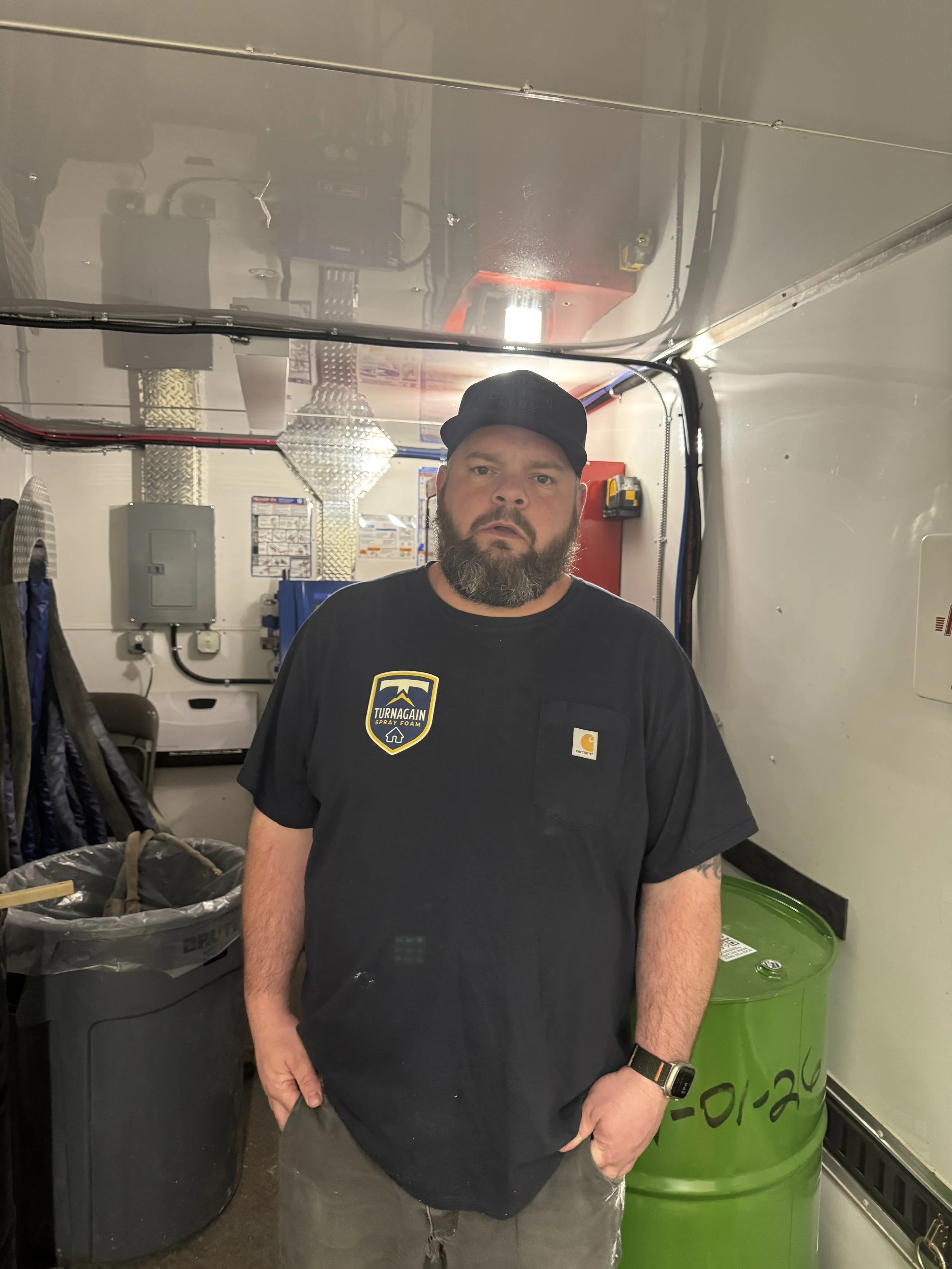 A man with a beard wearing a black cap and black t-shirt stands in an industrial or utility room, surrounded by various equipment and storage containers.