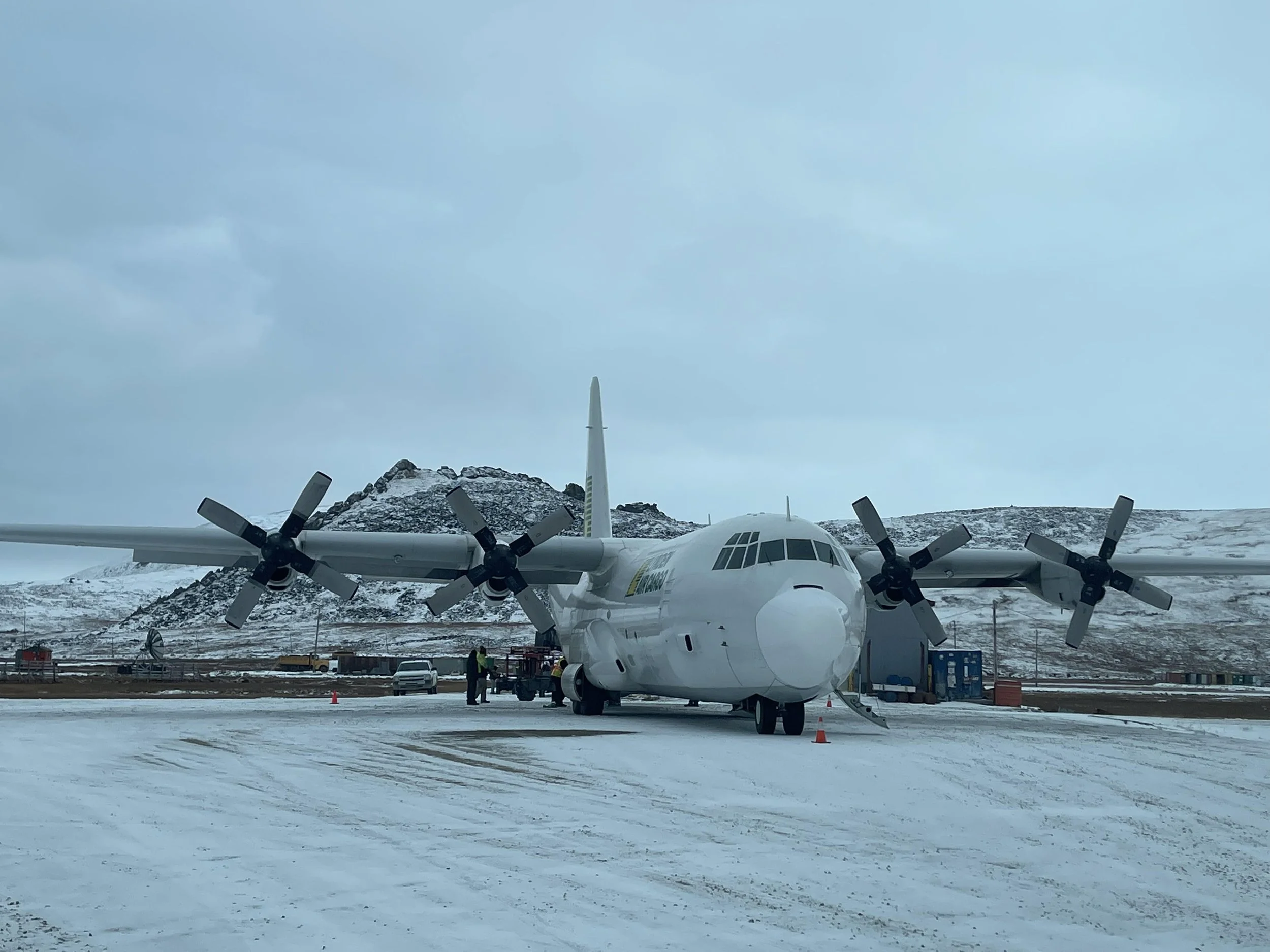 Large white airplane with four propellers parked on snow-covered ground at an airport, with mountains and a cloudy sky in the background.