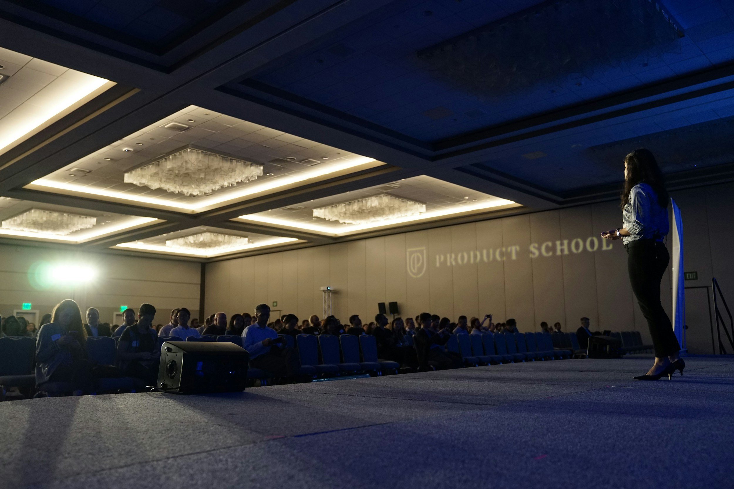 A woman giving a presentation on stage at a conference with an audience seated in front of her. The room has a high ceiling with decorative lighting, and a projection on the wall reads 'Product School'.