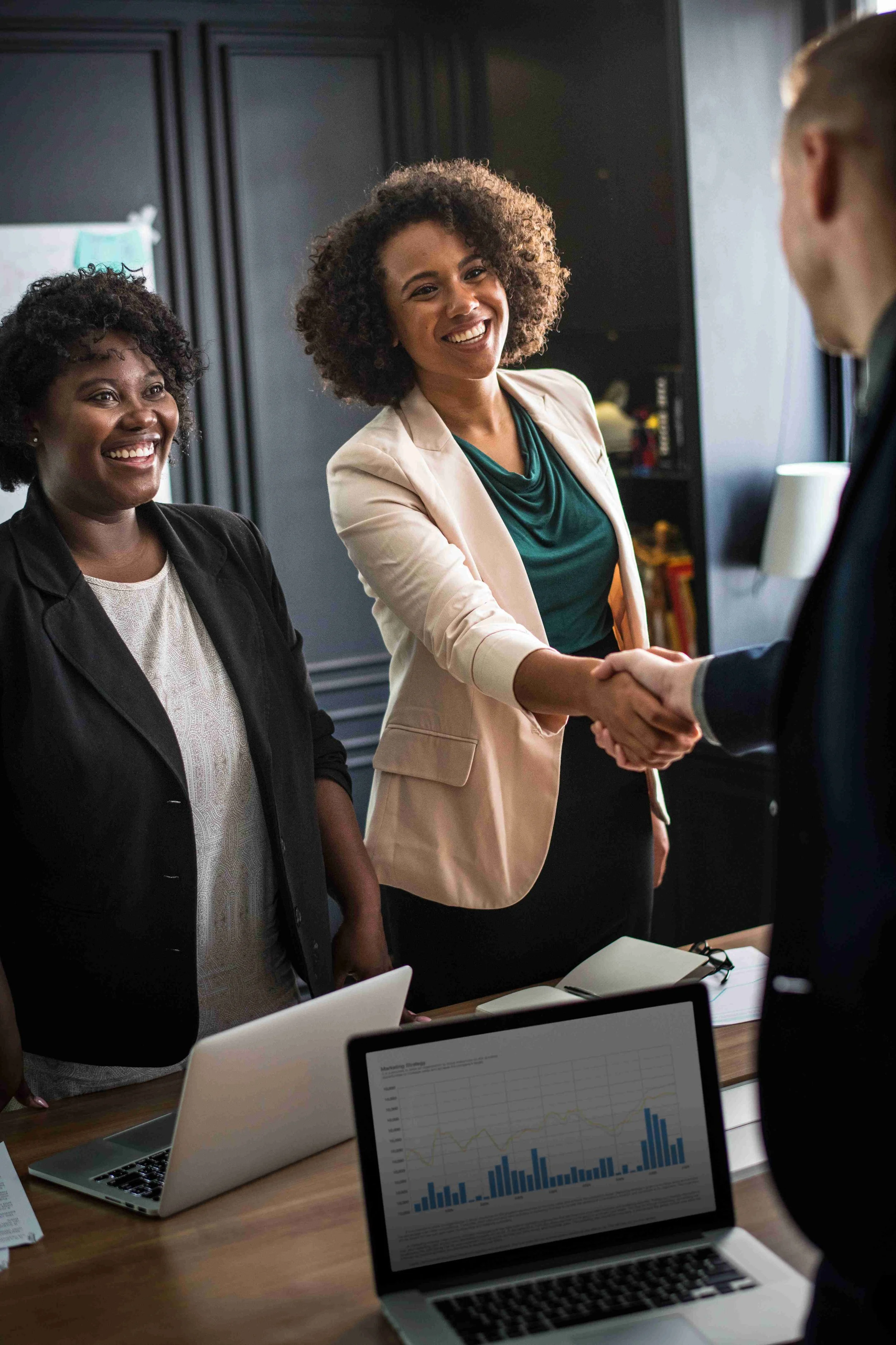 Two women in business attire shaking hands in a meeting room, smiling. A third woman stands beside them, also smiling. There are laptops on the table and a chart displayed on an open laptop in the foreground.
