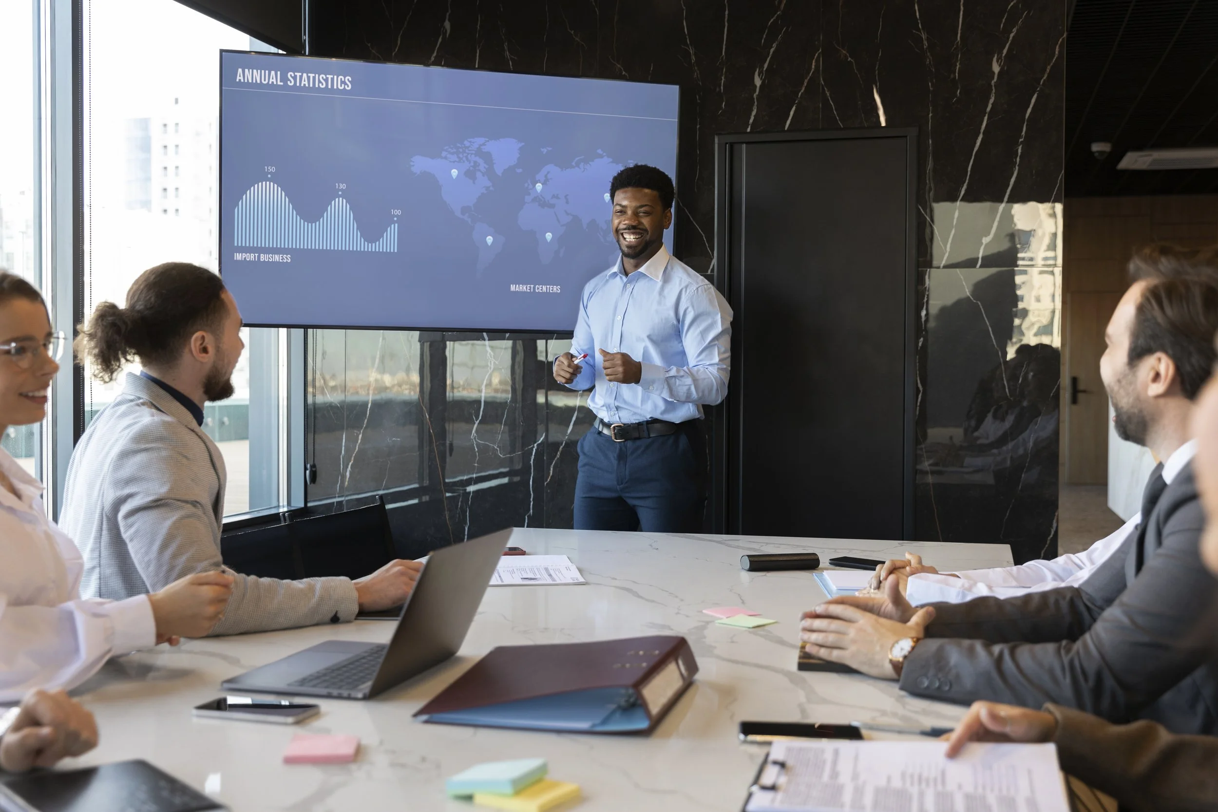 Man leading a presentation in a meeting room with colleagues, a large screen displaying 'Annual Statistics' with graphs and a world map.