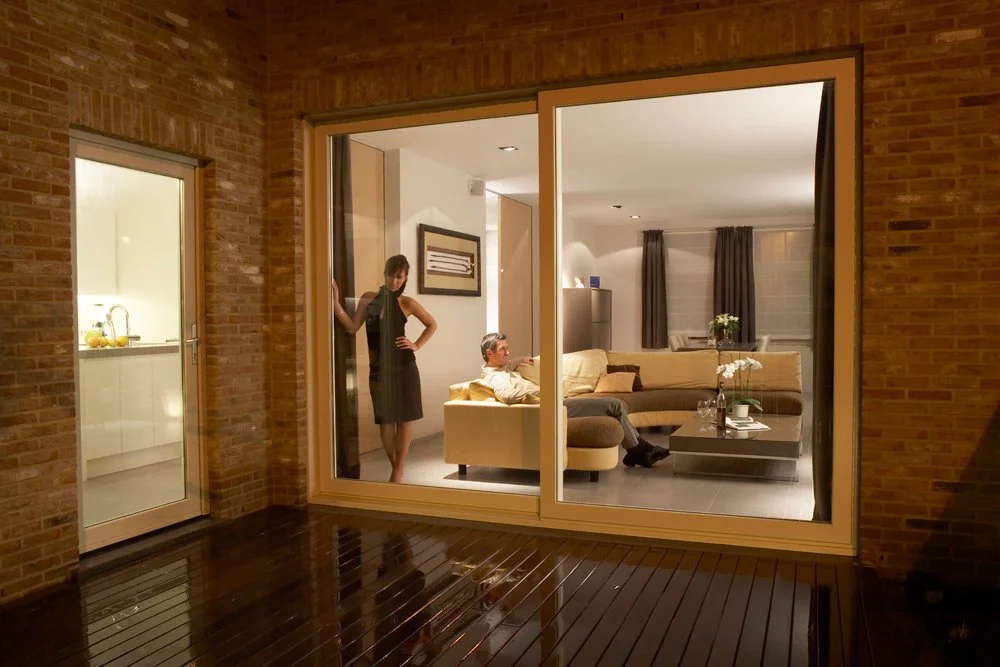View of a modern living room seen through large glass sliding doors from a wooden patio, with a woman standing inside near a wall and a man sitting on a beige sectional sofa.