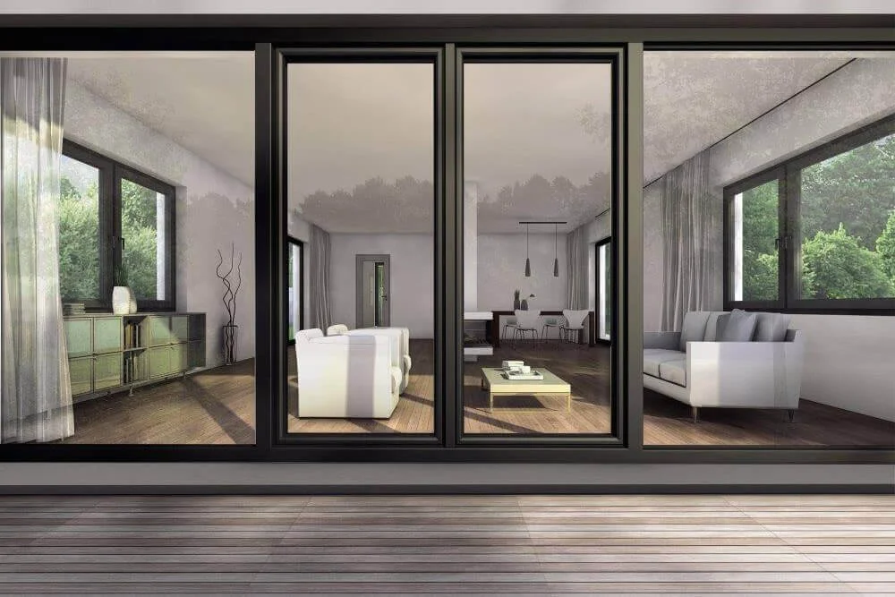 Interior view of a modern living room with large windows, white couches, and a dining area in the background, seen from an outdoor deck.