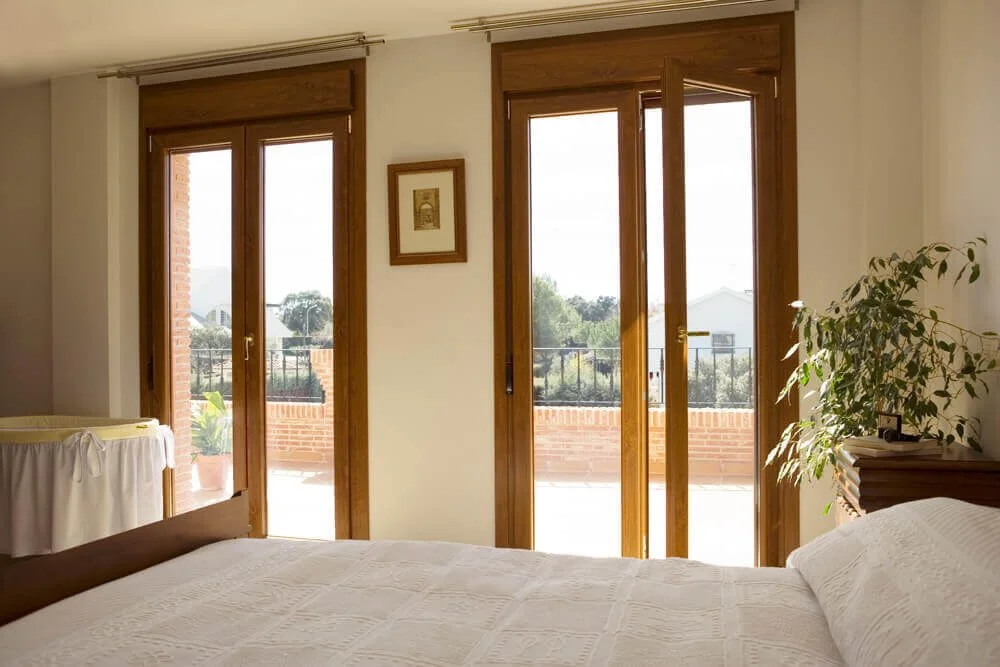 Bedroom with three large glass doors leading to a balcony, wooden frames, white bedspread, small framed picture on wall, potted plant on nightstand.