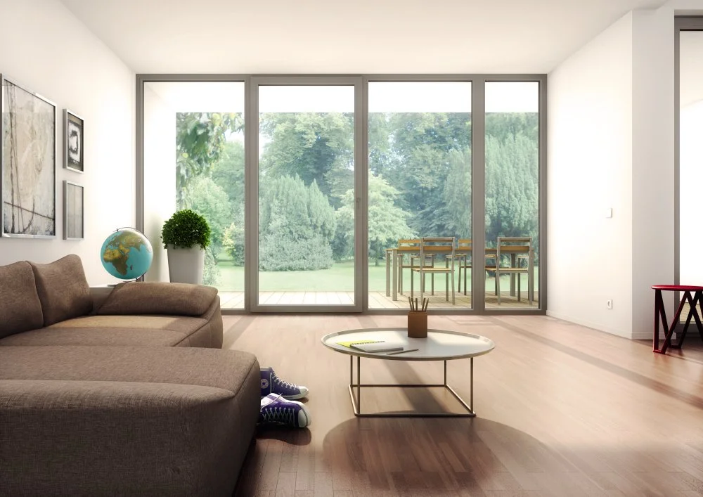 Modern living room with large glass sliding doors overlooking a green outdoor area. Features include a beige sofa, a globe, a potted plant, a white coffee table with books and a pencil holder, and framed artwork on the wall.