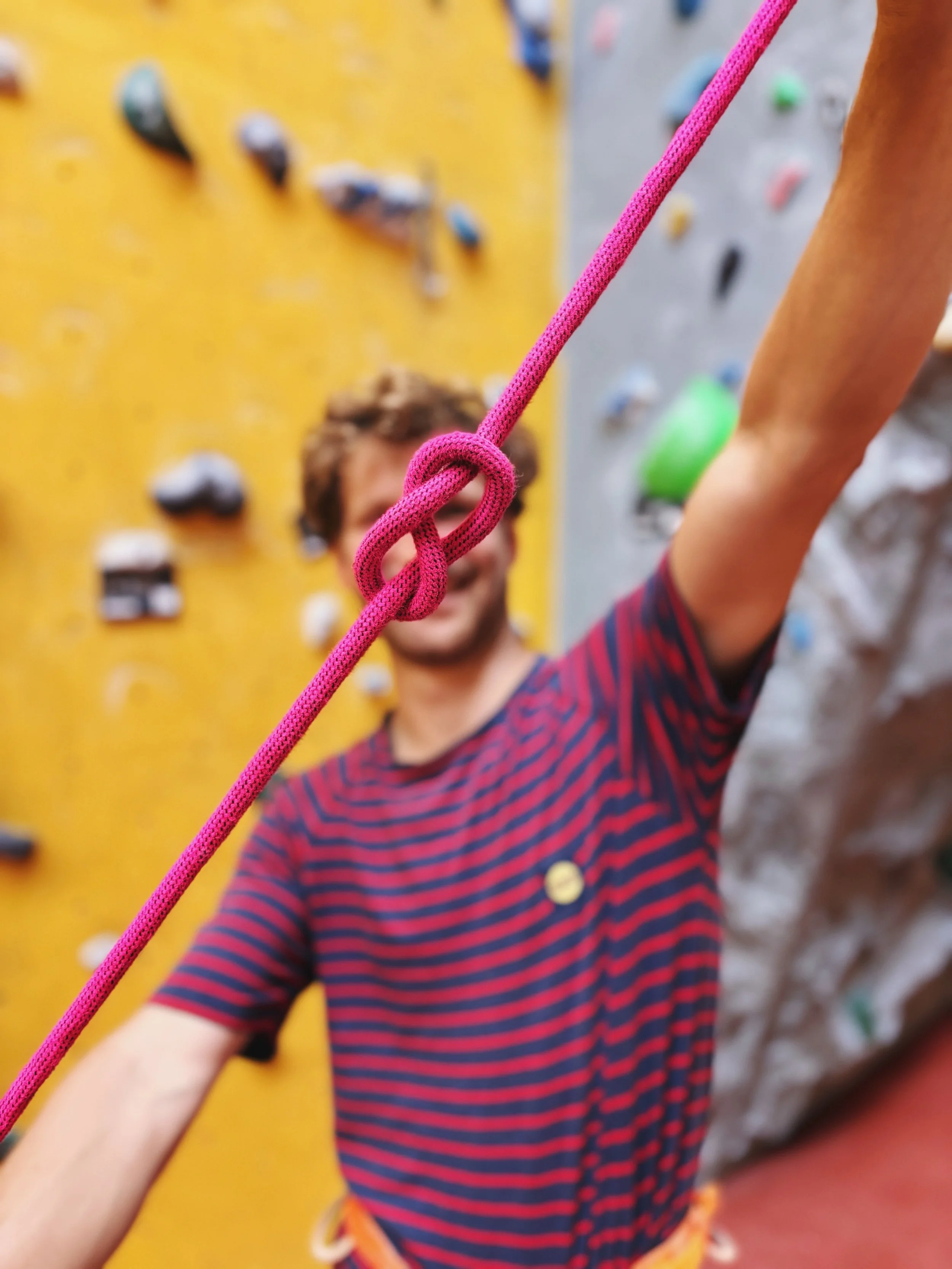 Person beim Klettern, Fokus auf Knoten in der Sicherheitsleine, im Hintergrund eine Kletterwand mit Griffen.