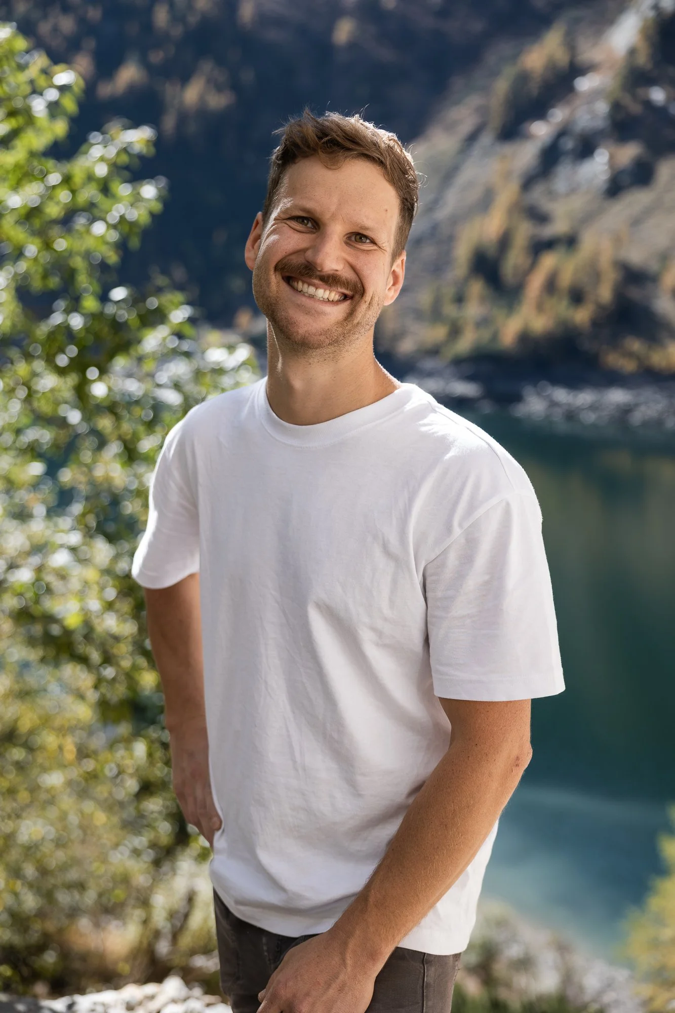 Ein lachender Mann mit kurzen braunen Haaren und Bart, trägt ein weißes T-Shirt, steht vor einer natürlichen Kulisse mit Wasser und Bergen im Hintergrund.