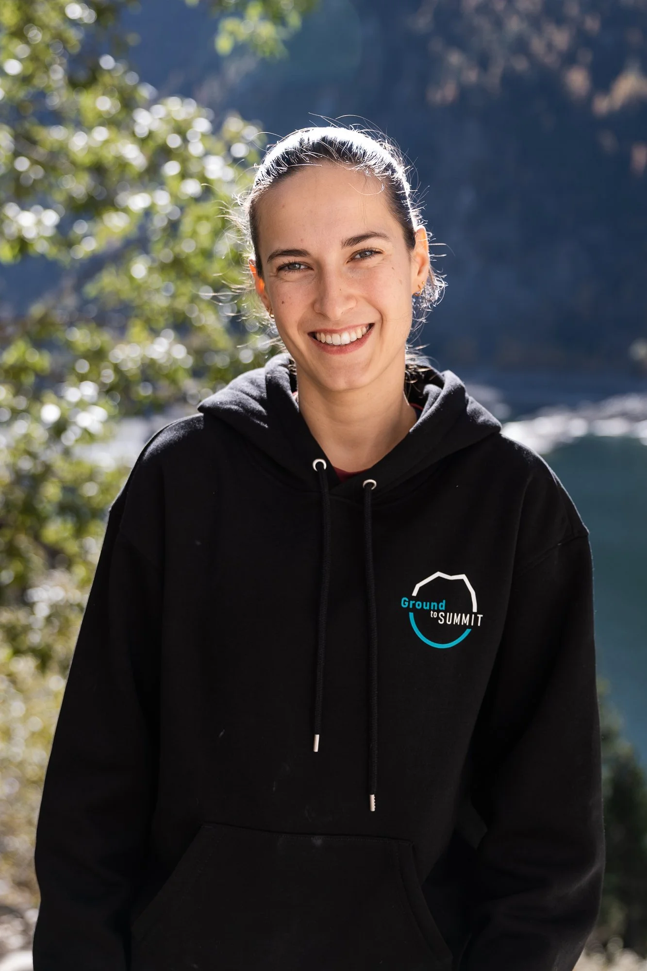 Junge Frau im schwarzen Hoodie mit Aufdruck 'Ground to Summit' vor einer natürlichen Landschaft mit Wasser und Bäumen
