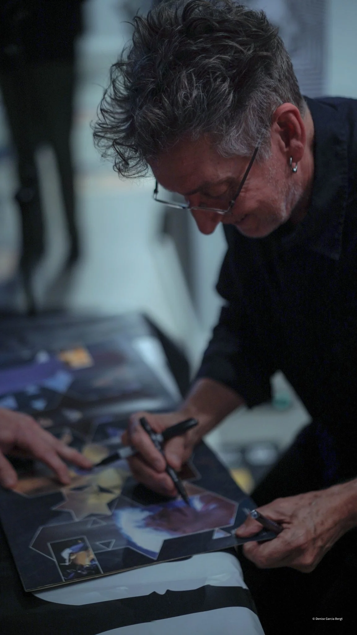 A man with gray hair, glasses, and earrings is signing a photograph or poster with a black marker, leaning over a table with multiple photos or posters on it.
