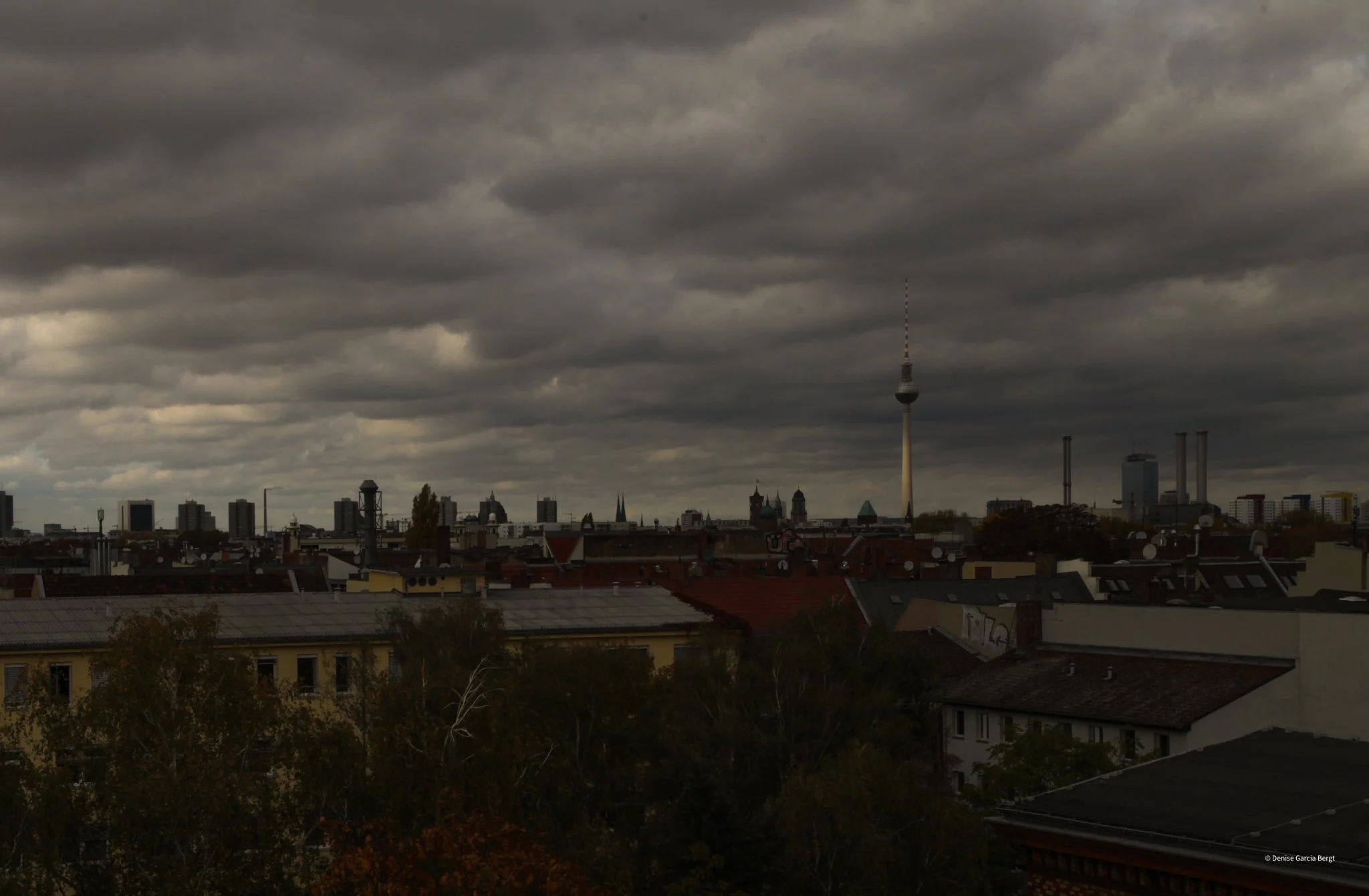 Overcast city skyline with dark clouds, showing a TV tower and various industrial and residential buildings.
