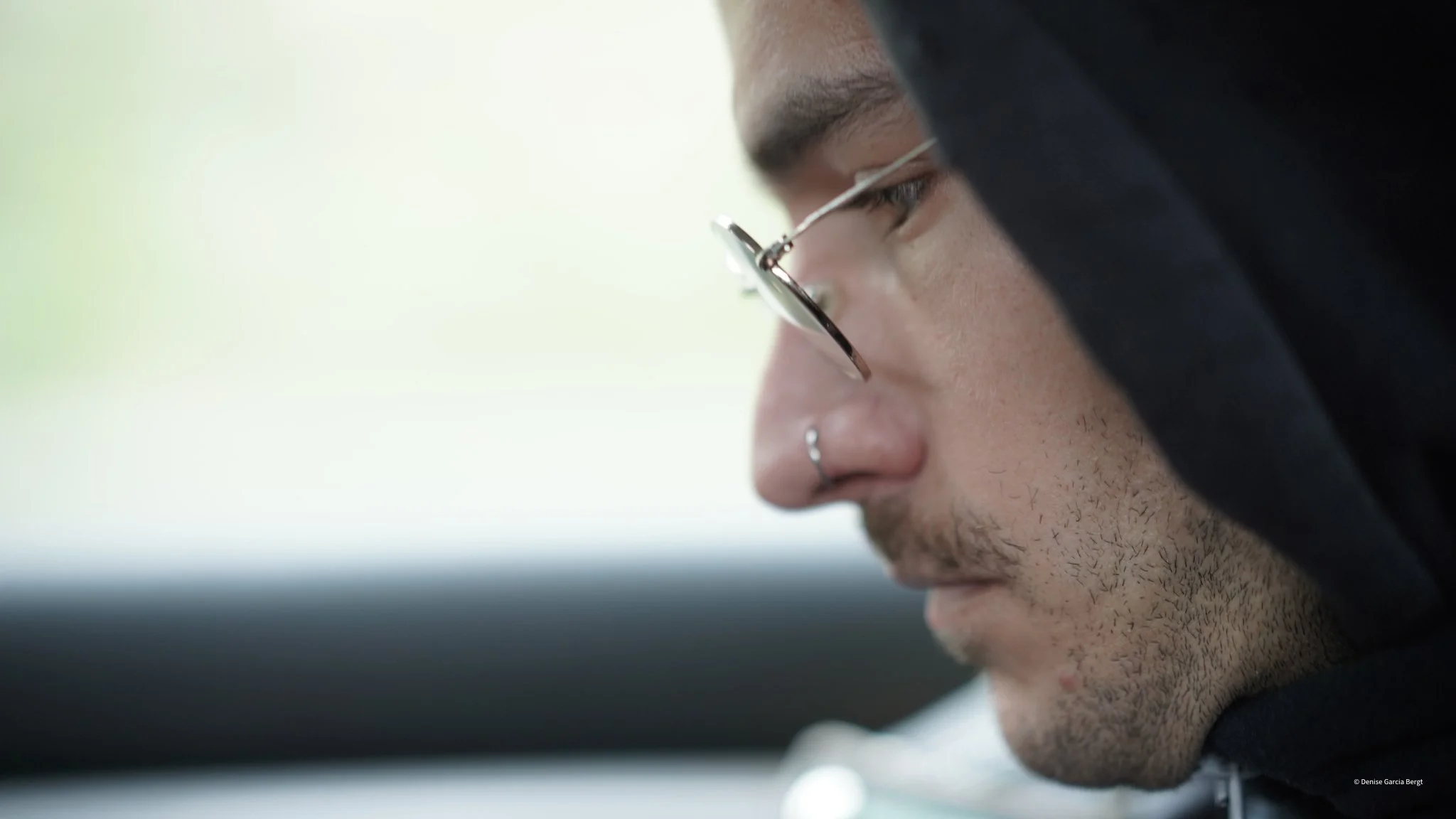 A close-up side profile of a young man with glasses and a nose piercing, wearing a black hoodie, looking down. The background is blurred.