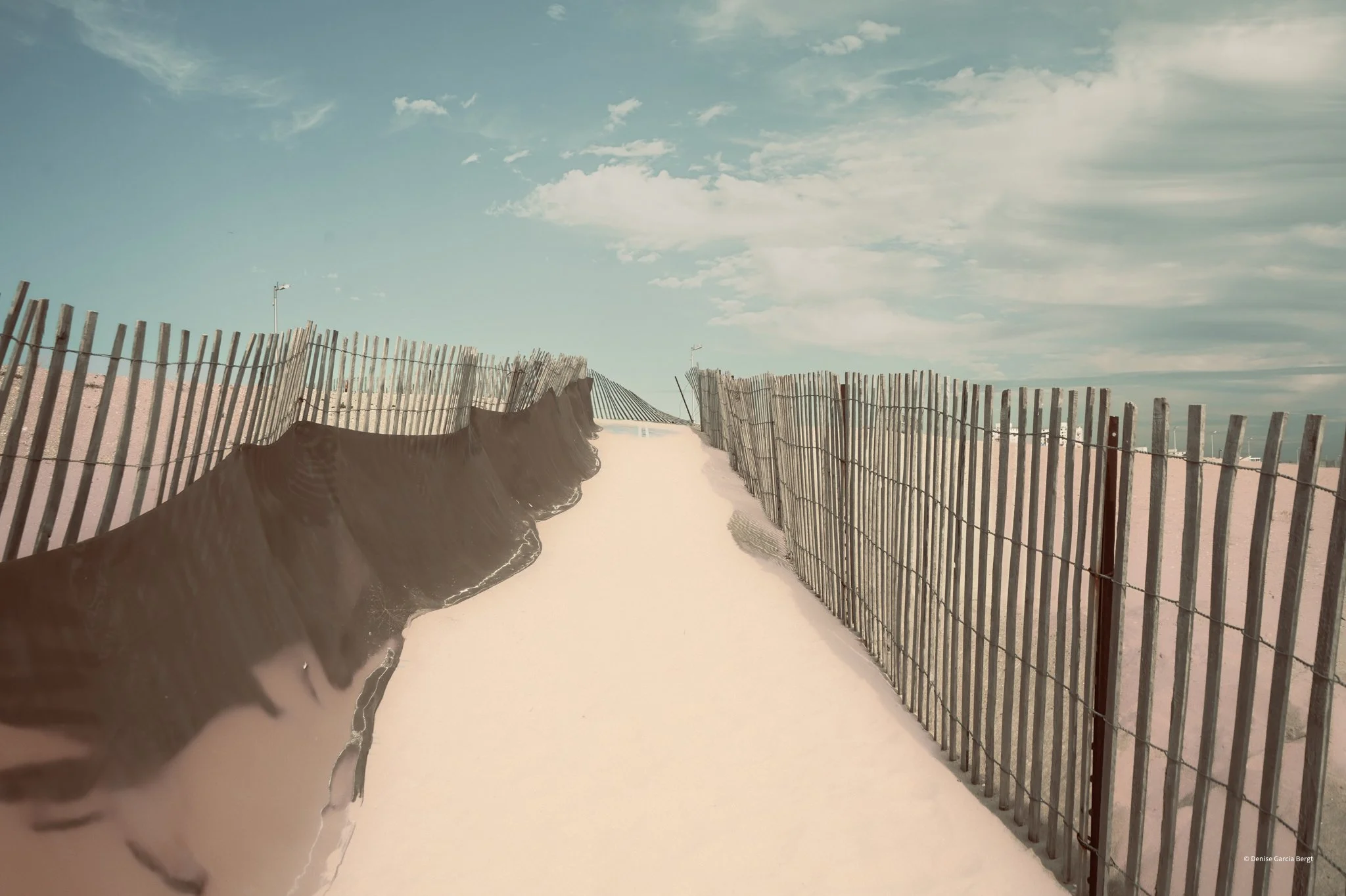 Pathway with tall wooden fences on both sides leading towards the horizon under a partly cloudy sky.